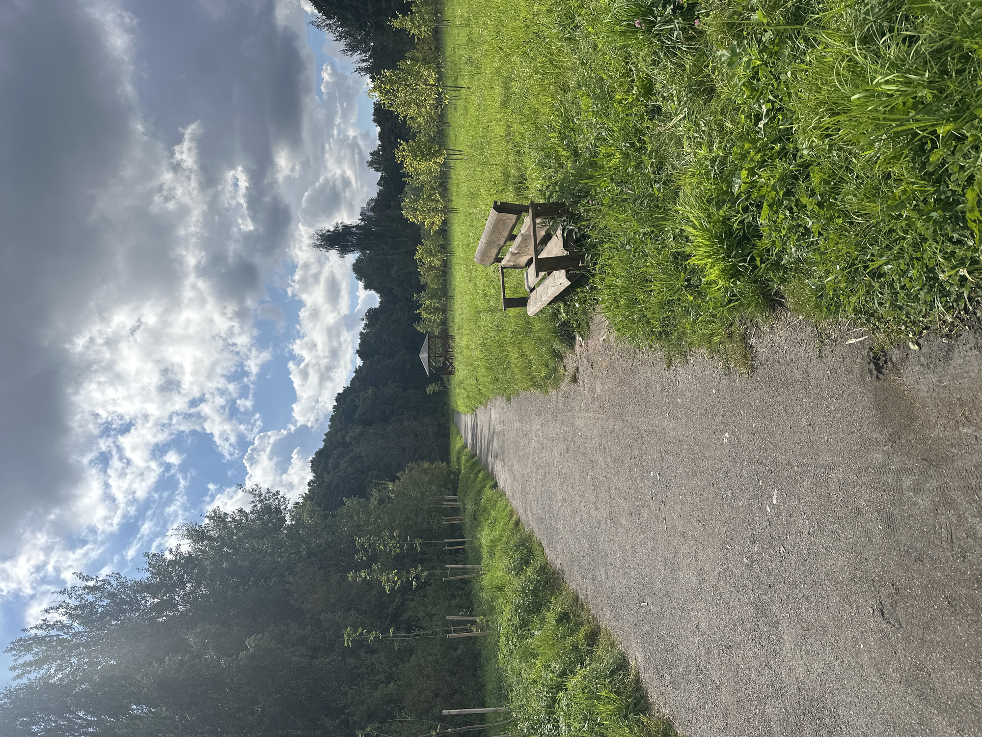 The image shows a gravel walking path in a park in Minsk, Belarus. The path curves to the right, leading into a grassy field dotted with young trees. A wooden bench sits to the right of the path, in the grassy area. In the distance, a small wooden gazebo is visible. The sky is partly cloudy, with dramatic clouds interspersed with patches of blue sky. The overall scene suggests a tranquil, natural setting within the city.