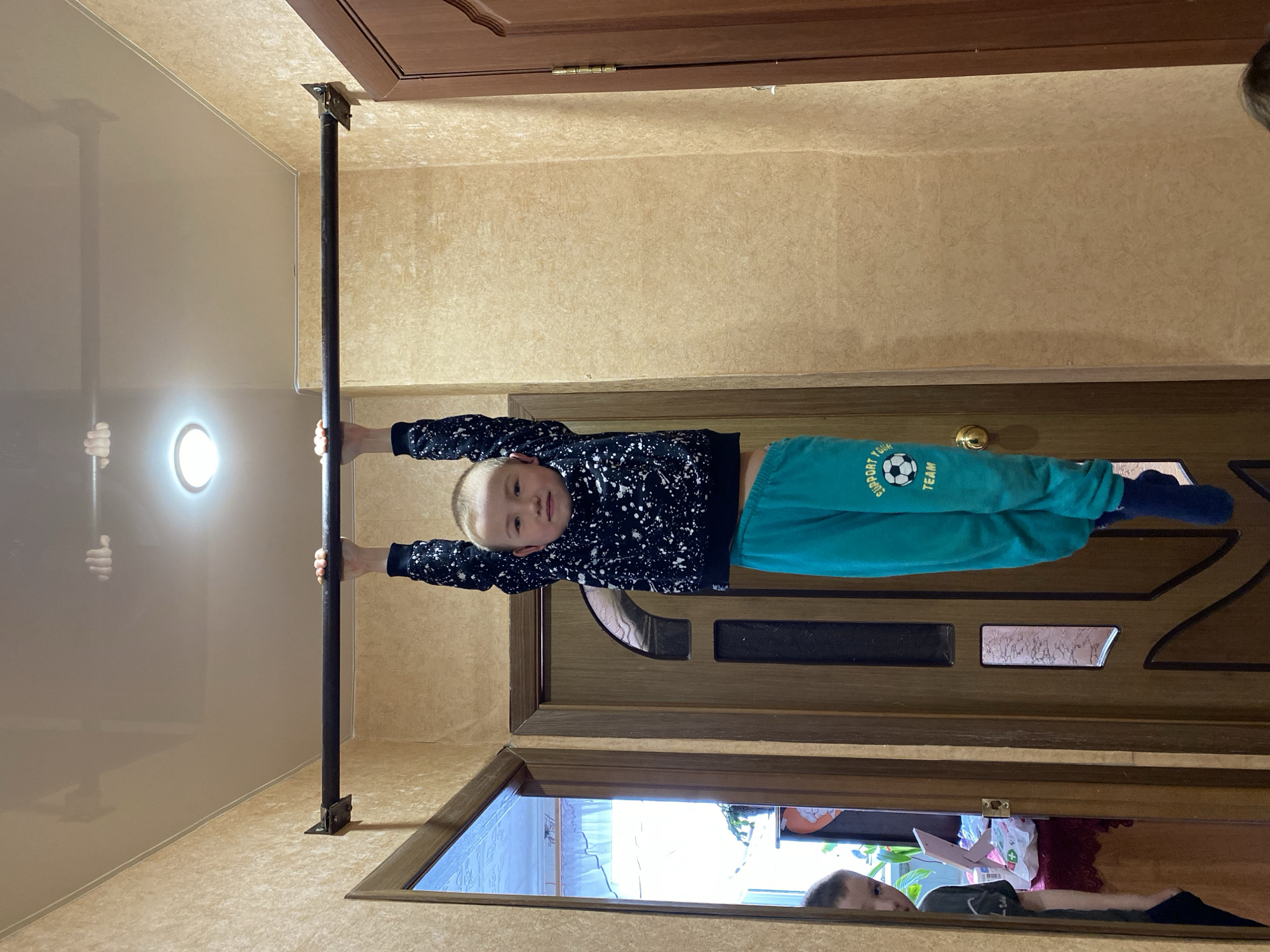 A boy is hanging from a metal bar installed in a doorway. He is wearing a dark patterned sweatshirt and green sweatpants. Another person is visible through the open doorway below, partially obscured by objects, but no other information about the location in Bely Klyuch, Russia, can be ascertained from the image.