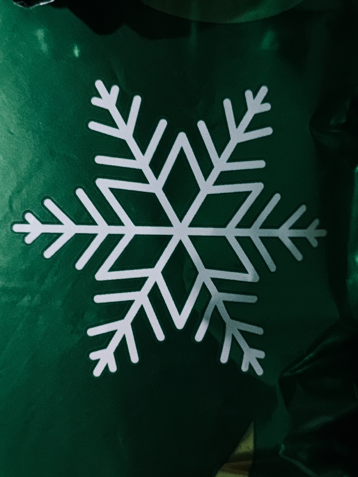 A close-up shot reveals a white snowflake centered on a background of emerald green material. The snowflake exhibits a symmetrical, six-pointed design with angular branches. The green surface appears to be glossy or reflective, creating soft highlights and shadows. A partial glimpse of a golden object is visible at the bottom, near the right edge of the image.