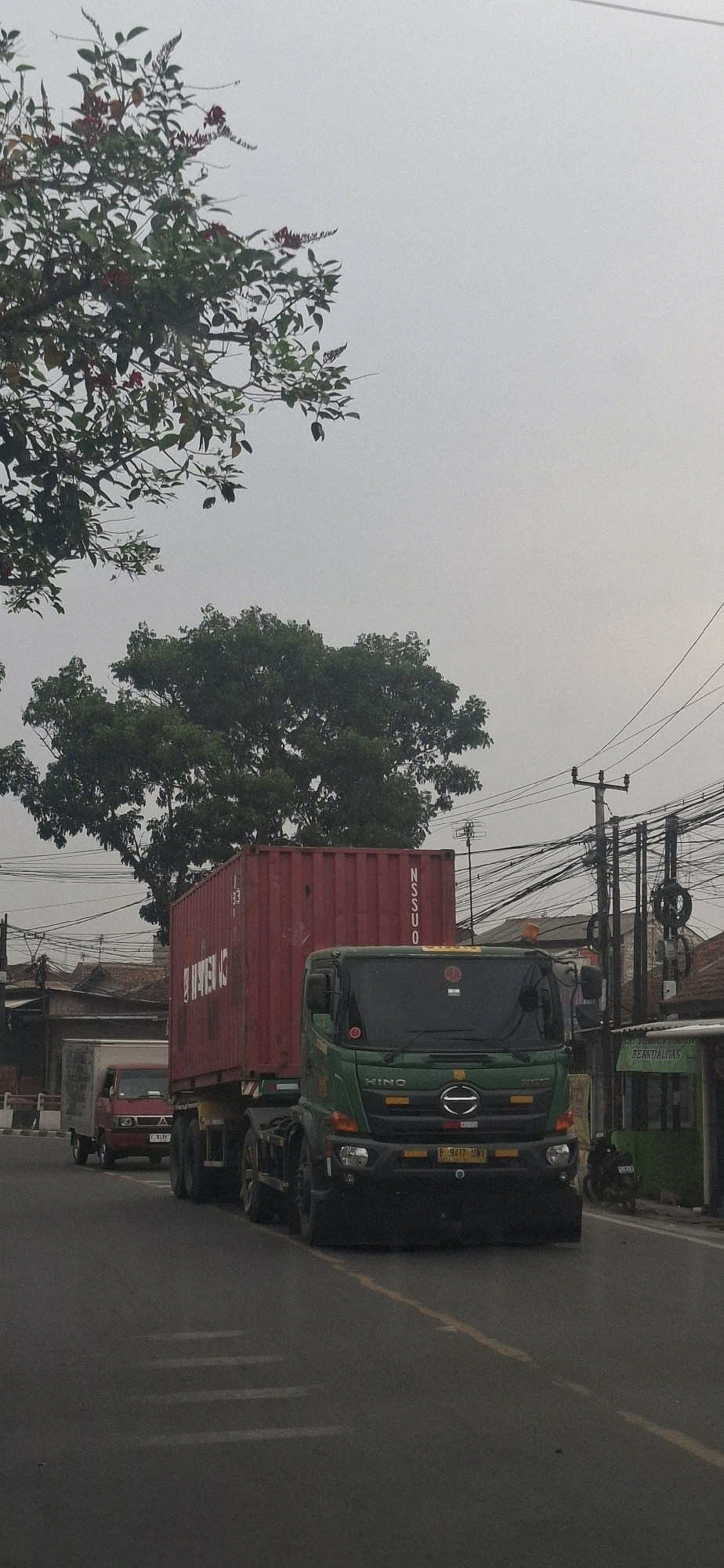 The image shows a street scene in Sirnagalih, Indonesia. A green truck with a red shipping container is on the right side of the road, in motion. To its left, a smaller red truck is also on the road, in front of the larger truck. Overhead, power lines and utility poles are visible, along with trees and vegetation. The sky is overcast. Buildings and structures line the right side of the street. A motorcycle is parked on the right side of the road near a building. The road surface is asphalt with painted lane markings.
