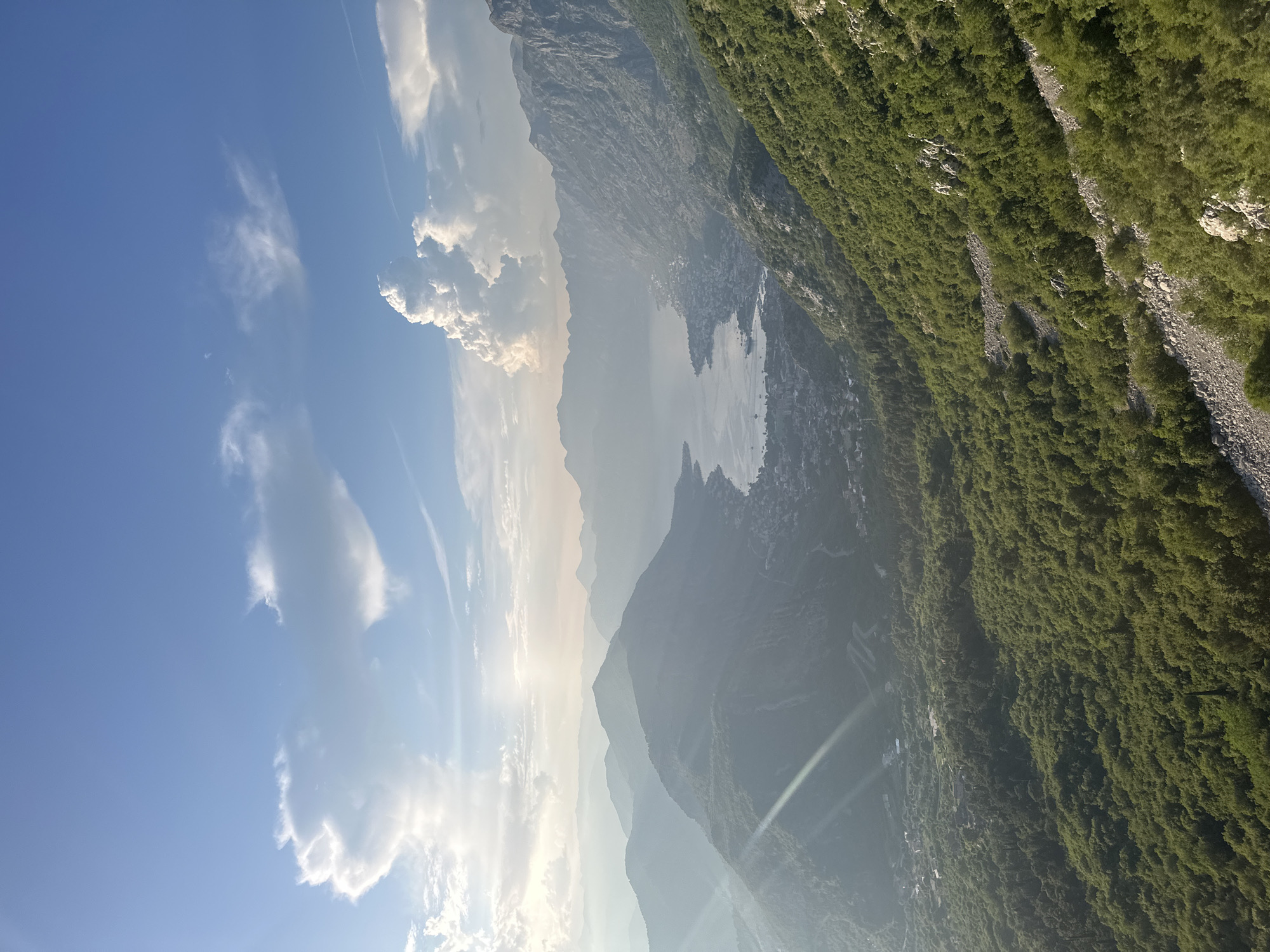 An aerial view captures a sunlit bay surrounded by lush green mountains, with a town nestled along the coastline. The sky is a bright blue, adorned with scattered white clouds. The foreground features a steep, tree-covered slope, with a rocky path winding down it. In the midground, a body of water is visible, with a town spread out along its edge. Mountains rise in the background, their peaks fading into a hazy distance. The image appears to be taken from a high vantage point, offering a panoramic perspective of the landscape. The scene is characteristic of the Bay of Kotor, with the town visible potentially being Škaljari or a neighboring settlement in Montenegro.