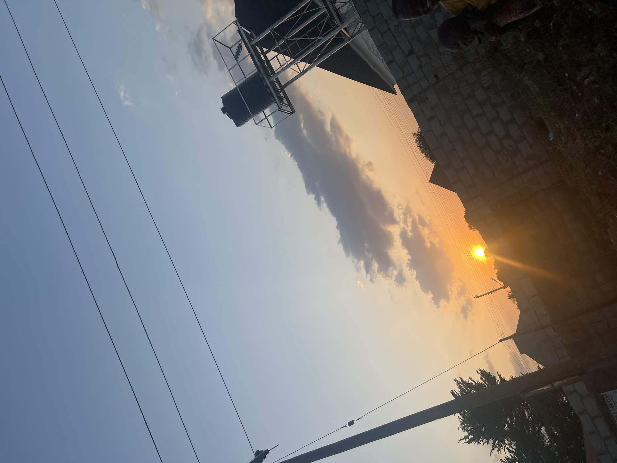 The image depicts a sunset scene in Abuja, Nigeria. The sky is a mix of blue and orange hues with clouds obscuring the sun. Foreground objects include power lines and a metal structure with a water tank. The edge of a building is visible, including a paved area, along with some shrubbery. Two people are standing on the paved area.