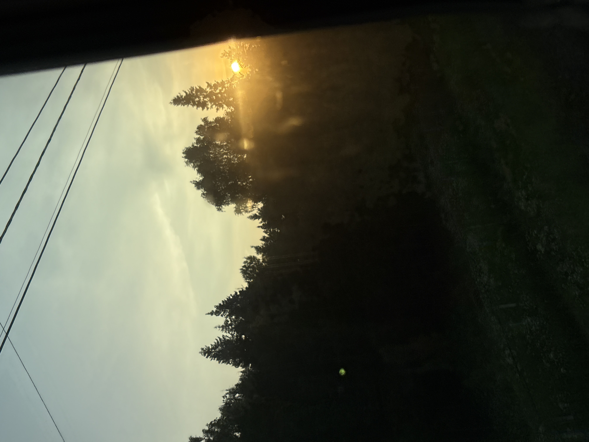 The image captures a sunset viewed through a window, likely from a moving vehicle. The foreground is dominated by a dark silhouetted treeline against a sky transitioning from a pale yellow at the horizon to a light blue at the top. The sun, a bright orange orb, is partially obscured by the trees, casting a warm glow and creating sunbeams that pierce through the foliage. Several dark, thin power lines are visible stretching across the upper portion of the frame. The overall scene suggests a natural landscape at dusk, possibly on the outskirts of a city.