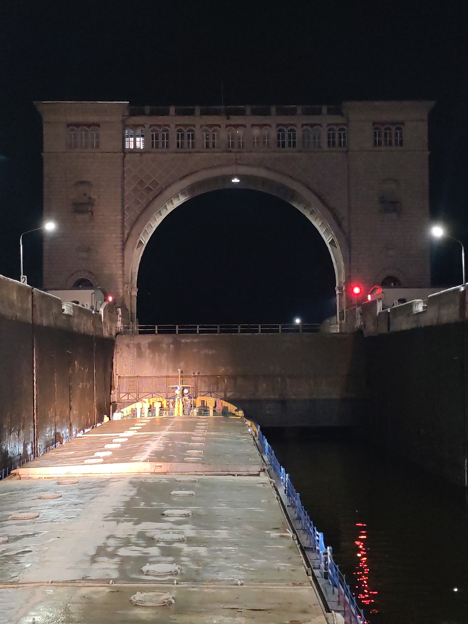 The image shows a large, illuminated lock gate structure at night, located in the city of Vasil'ki, Russia. The structure features a massive arched gateway made of stone, with several windows on its upper facade. Two lampposts are visible, one on the left and one on the right, illuminating the scene. A red traffic light is also visible on the left side of the structure. In the foreground, a wide, wet ramp or platform extends into the lock, with several circular manhole covers or access points visible on its surface. The water within the lock is dark, with reflections of red lights visible on its surface. The overall impression is one of an industrial or infrastructural site at rest, under artificial light in the darkness. There are no people visible in the image.