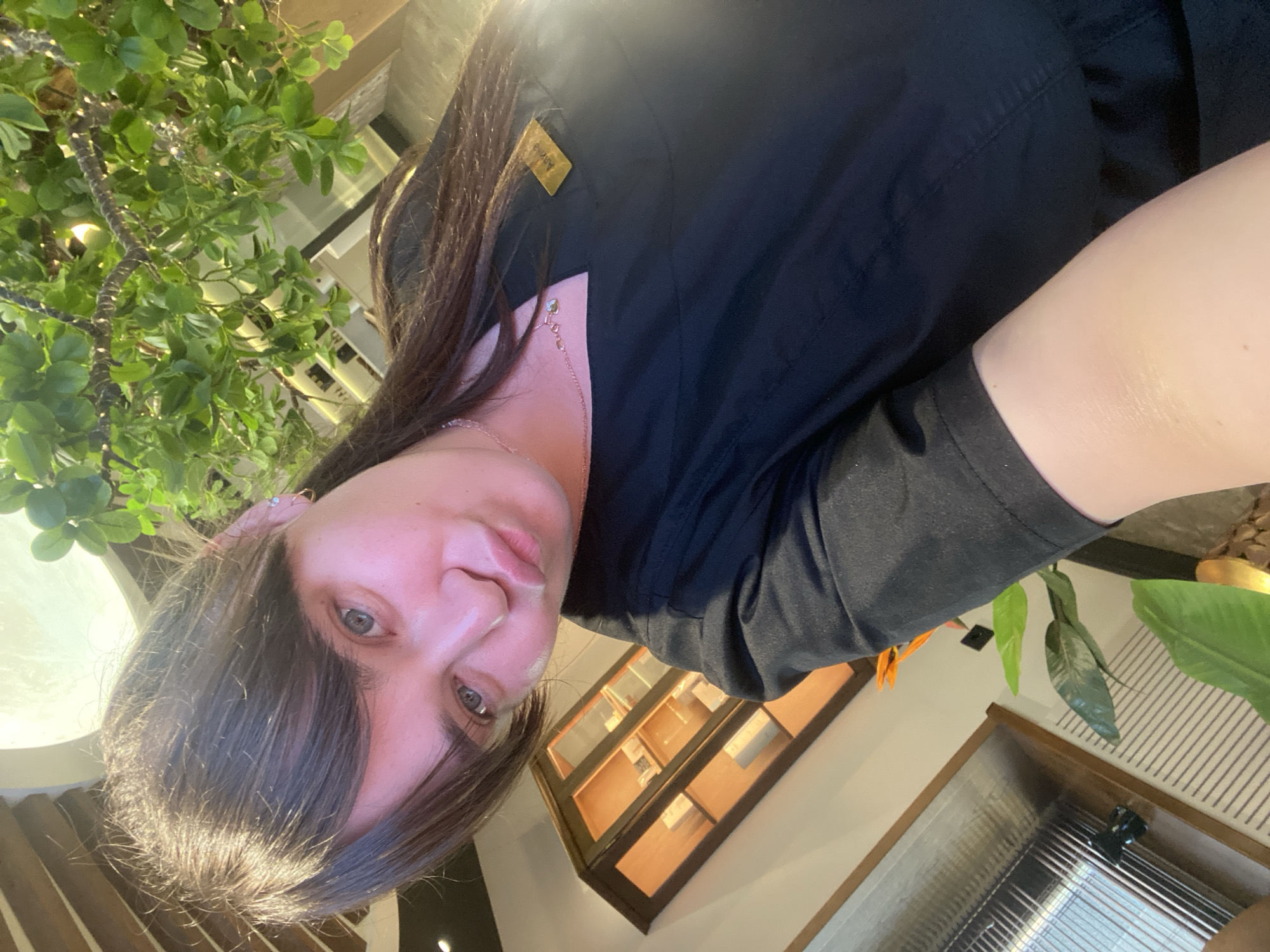 A selfie taken by a woman with dark hair and blue eyes, wearing a black garment and a delicate silver necklace. She is indoors near decorative plants, wooden shelving, and a metallic container. The location is presumed to be Votkinsk, Russia.