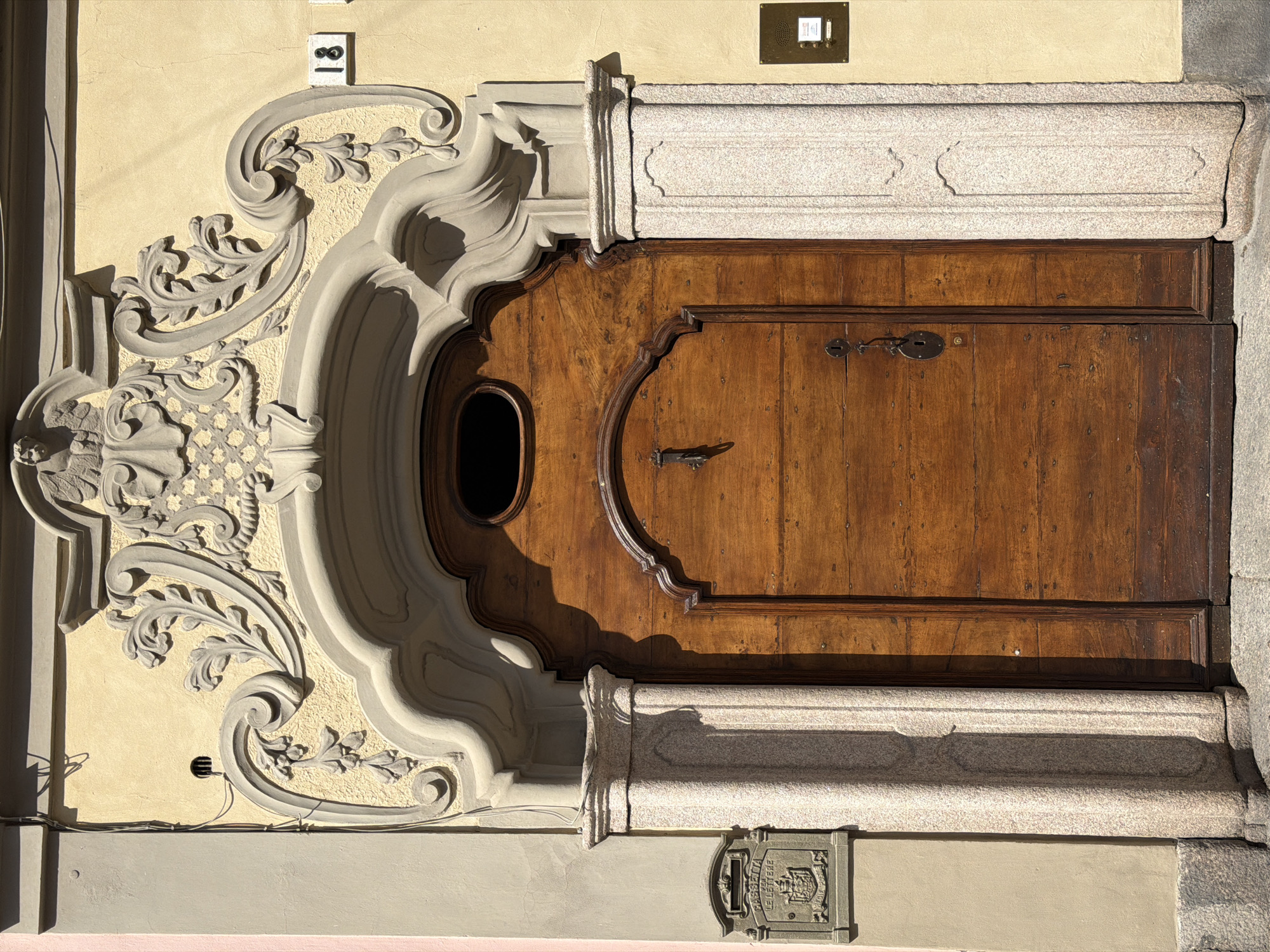 This is a vertical photograph showcasing an ornate wooden entrance door set into a light yellow stucco wall, characteristic of an old building facade in Vigevano, Italy. The dark brown wooden door features rich graining, detailed paneling, and a distinct oval-shaped window in its upper left section. Traditional metal hardware, including two keyhole escutcheons and a decorative pull handle, are visible on the door's surface.
The door is enveloped by elaborate architectural details. On the left, a grand stucco or carved stone surround displays complex Baroque-style floral and scroll ornamentation, casting deep shadows that emphasize its three-dimensional form. This intricate design sweeps from the upper door frame downwards. The door is also framed by robust granite pillars and a matching lintel above, which features subtle decorative carvings.
Several functional elements are integrated into the facade. A small white plaque with the black numbers "18" is affixed to the wall above the stucco ornamentation on the upper left. An antique-style brass intercom system, marked with the numbers "1" and "2", is mounted on the wall above the door on the right. Below the door, to the right, a prominent metal letterbox is embedded into the wall, clearly inscribed with the Italian phrase "CASSETTA DELLE LETTERE" and an ornate crest or coat of arms.
The scene is illuminated by bright, directional sunlight, creating sharp shadows that accentuate the textures and depth of the ornamentation and the door, suggesting a clear day, likely in the morning or late afternoon. A few minor wires run along the lower left portion of the wall. The image captures a sense of historical elegance and meticulous craftsmanship.