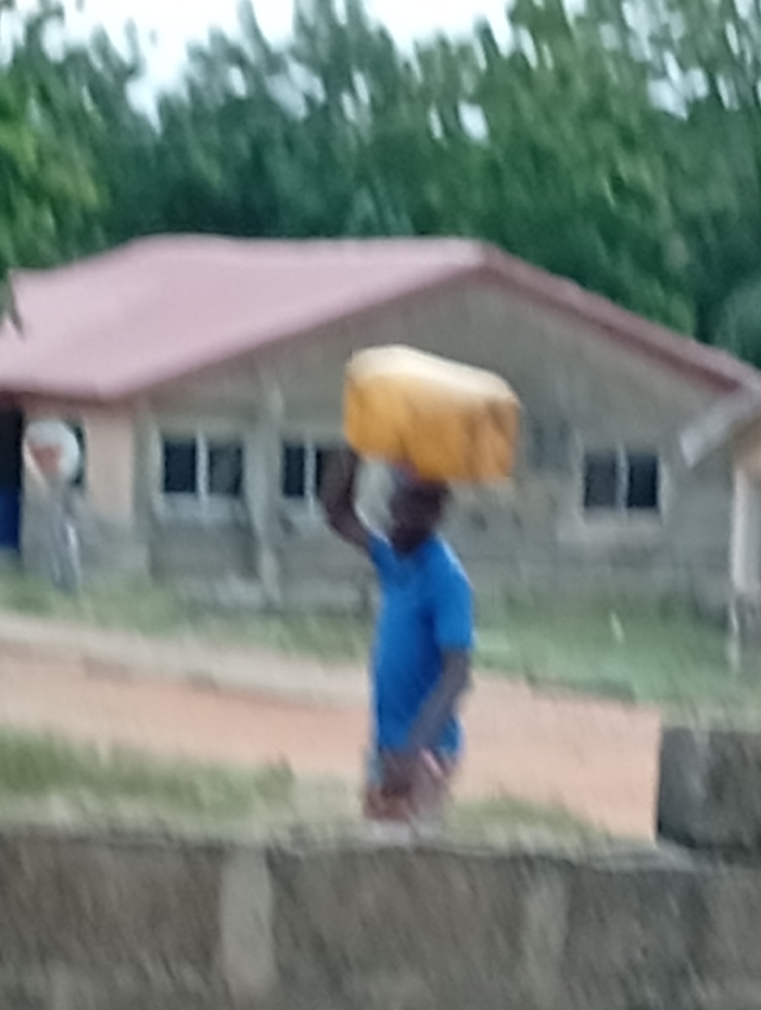 A blurred, low-resolution image depicts a man in Adankolo, Nigeria, carrying a yellow container on his head. He is wearing a blue t-shirt. Behind him is a one-story building with a red roof and several windows. The environment includes green trees in the background and a rough dirt path. A concrete wall is partially visible in the foreground. The scene suggests a residential or semi-rural area.