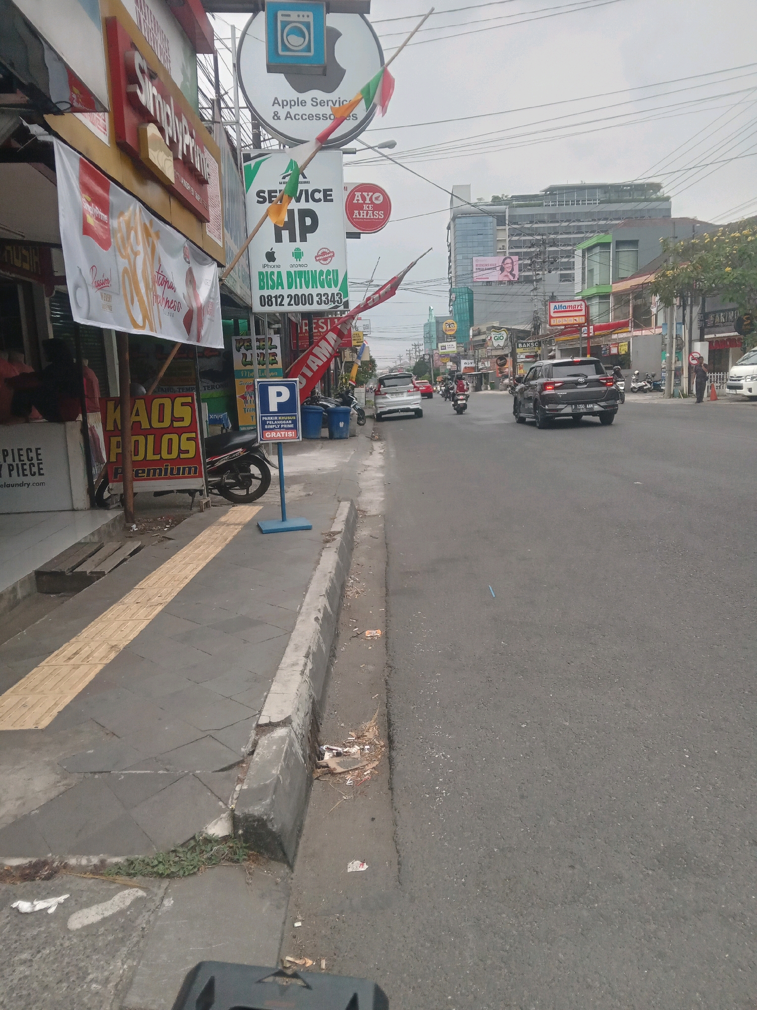 The image captures a street scene in Depok, Indonesia, featuring a bustling commercial area along a roadside. On the left side of the street, several shops are visible, including one with a prominent banner advertising "Simply Printing" and another displaying "KAOS POLOS Premium" (plain t-shirts). A parking sign for "SIMPLY PRIME" with the word "GRATIS!" (free) is also present. A motorcycle is parked near the shops. The sidewalk has textured yellow tactile paving.
Across the street, the road is busy with vehicles including cars and motorcycles. A dark-colored SUV is prominent in the foreground. Further down the road, buildings of varying sizes are visible, including a modern multi-story building with a green facade and a larger structure behind it that appears to be a commercial or residential complex with reflective windows. A sign for "Alfamart," a convenience store chain, is visible. In the distance, more traffic and buildings indicate a densely populated urban environment. The sky is overcast, suggesting a cloudy day. A person in dark clothing is standing on the right side of the road near a parked car.