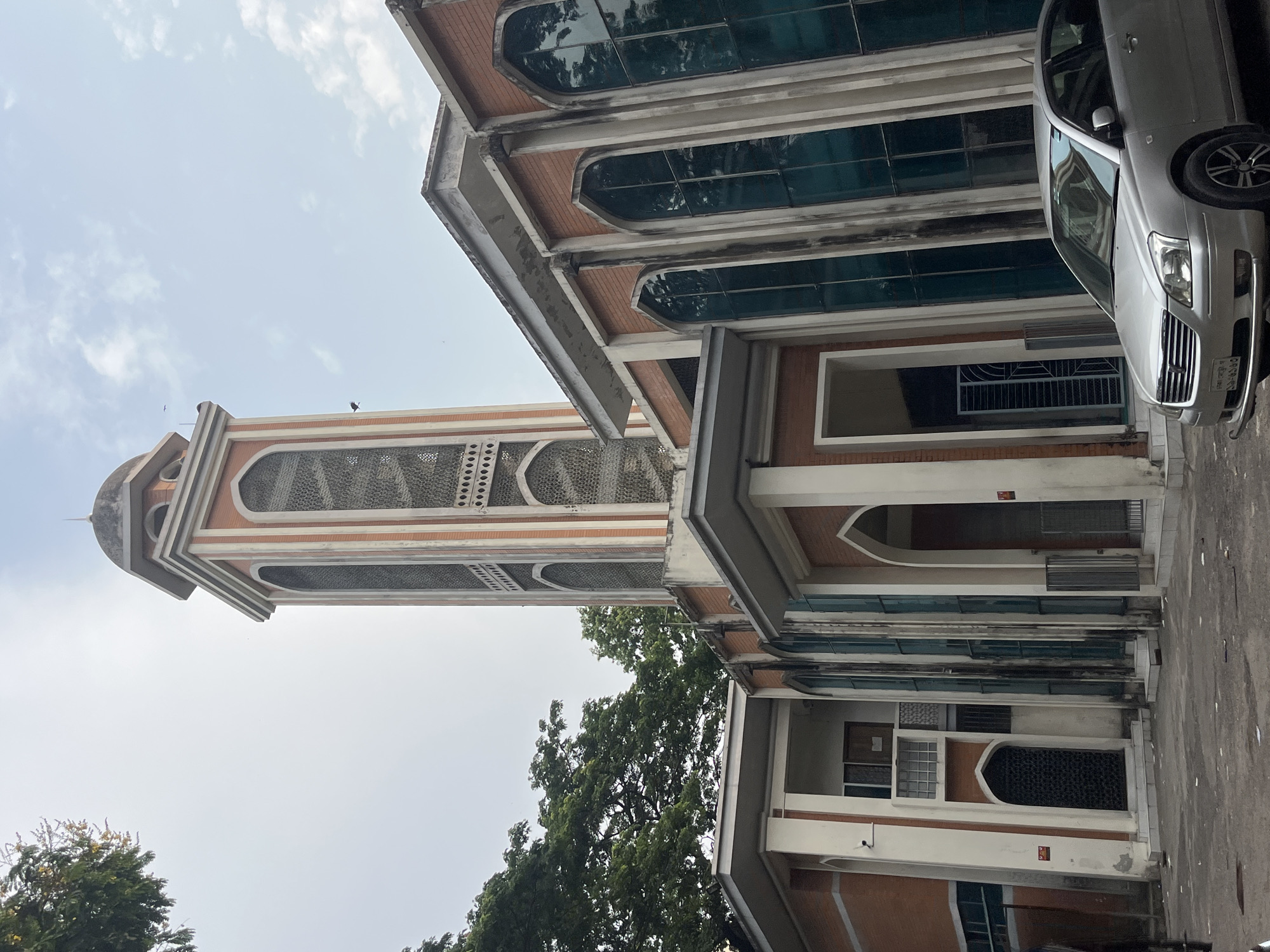 The image shows a multi-story building with Islamic architectural elements, including arched windows and a minaret. A silver car is parked in front of the building. The location is Dhaka, Bangladesh. The environment appears urban.