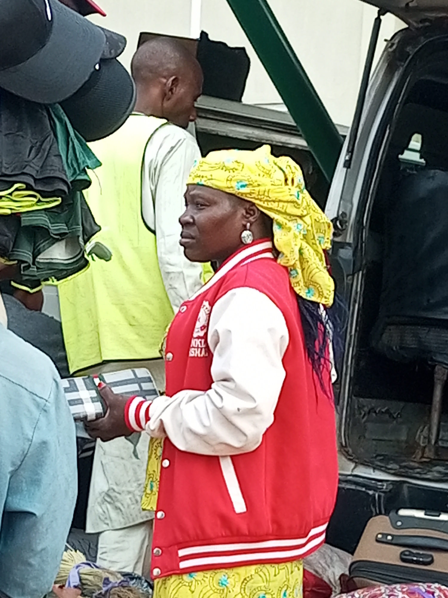 In Gombe, Nigeria, at what appears to be a roadside market, a woman wearing a yellow headscarf and a red and white bomber jacket is holding a checkered item. Beside her, a man in a neon yellow vest stands next to a vehicle with its trunk open. The scene is filled with various items, including hanging hats and stacks of folded clothing, suggesting a busy commercial area. The lighting indicates it is daytime.