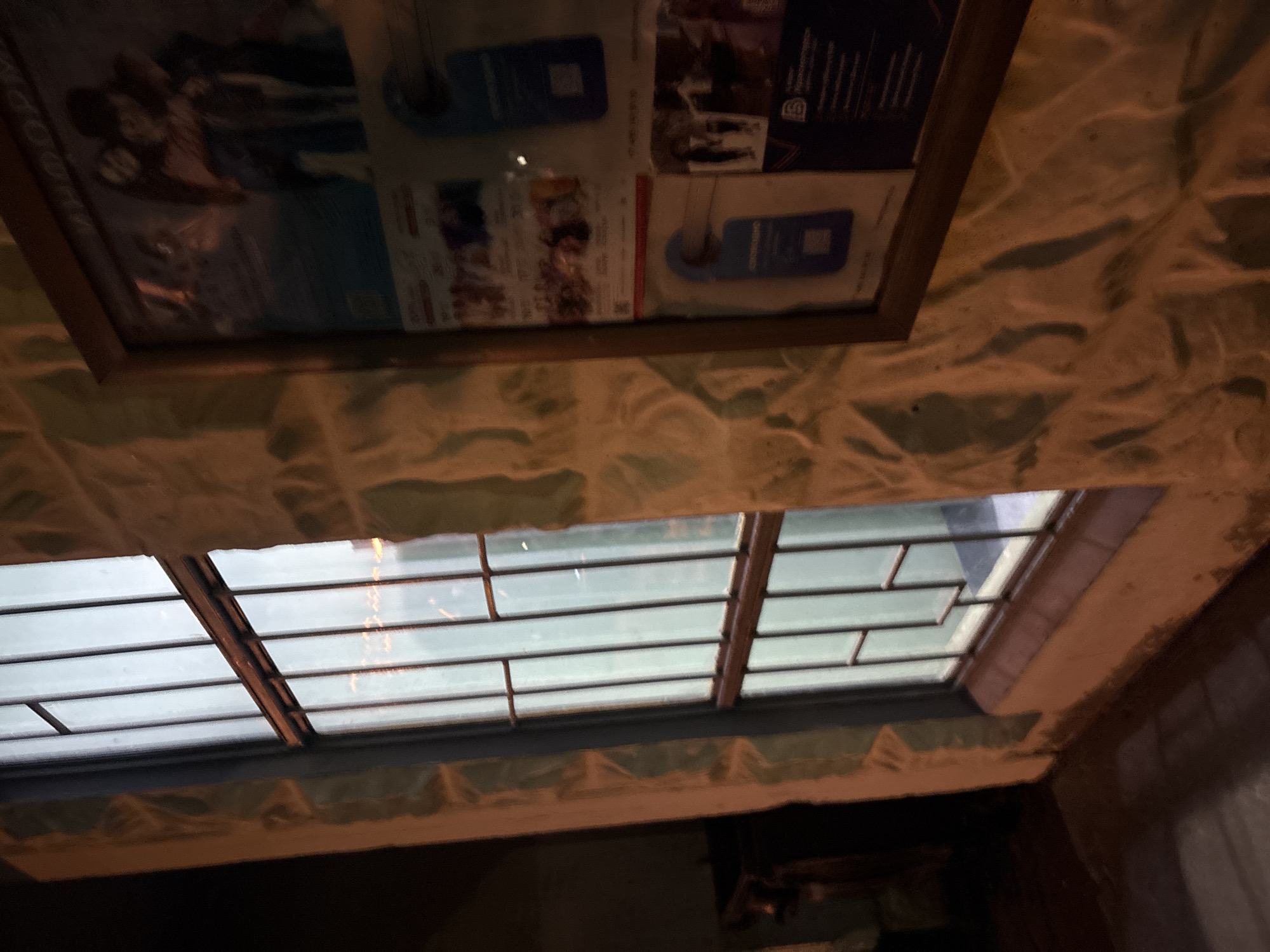 The image shows a window with metal bars, likely in Krasnogorsk, Russia. The window looks out onto a dimly lit area, possibly with some lights reflecting in the glass. To the right of the window, a framed bulletin board is visible, displaying several notices and possibly event posters. The wall is textured with a rough, stone-like finish. The overall lighting suggests it is either dusk or nighttime, with artificial light illuminating the scene.