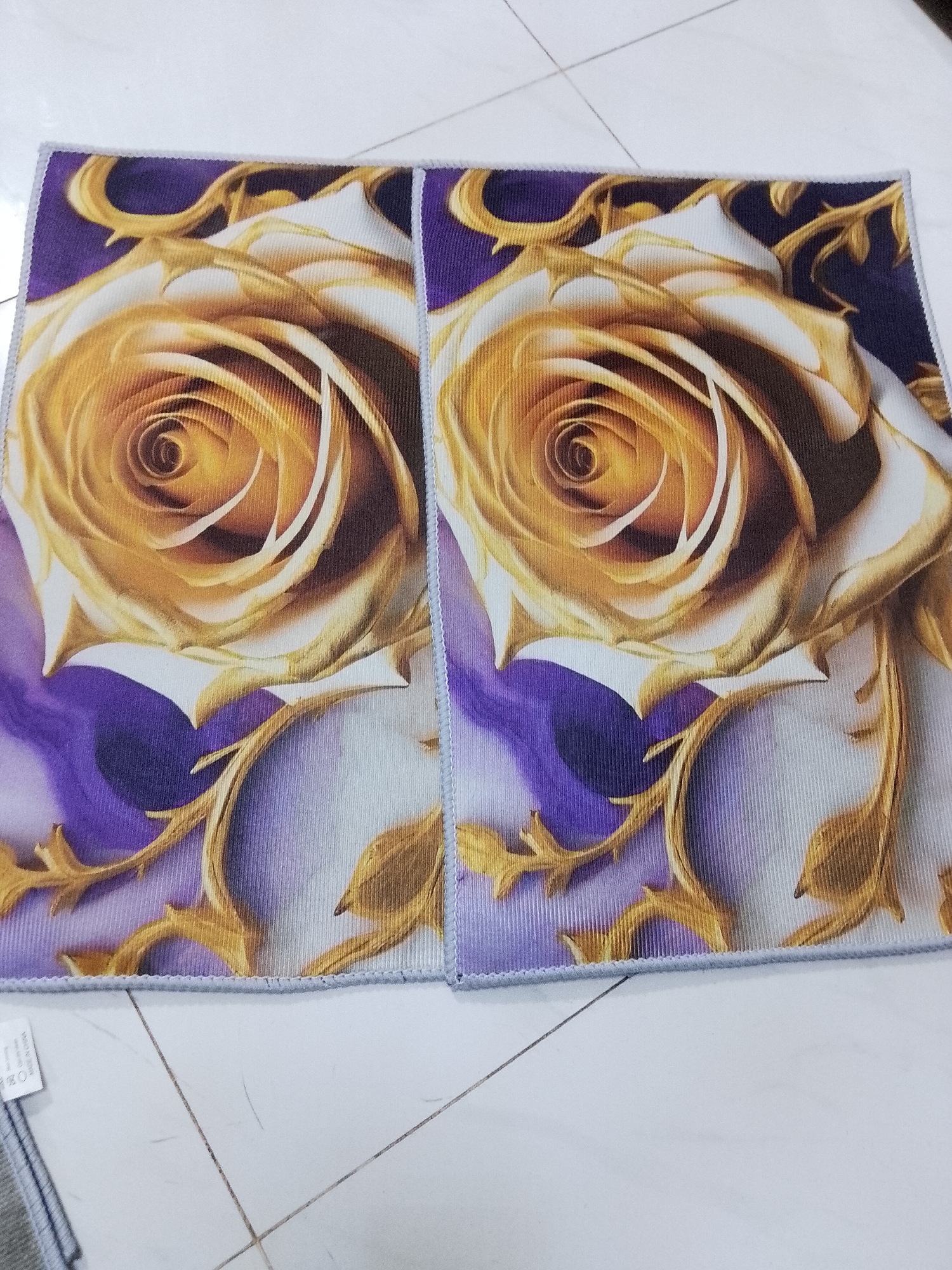 The image captures two identical rectangular fabric items, likely microfiber towels or small mats, displayed side-by-side on a light-colored tiled floor. The setting is an indoor space in Davao City, Philippines.
Each fabric piece features a striking design dominated by a large, intricately detailed rose. The rose petals are a rich golden-yellow in the center, transitioning to white on the outer edges, with dark brown shading providing depth. The background for the rose shifts from a vibrant, deep purple on the left to a softer lavender or light purple towards the right. Golden, swirling vine-like motifs are integrated into the background design, enhancing the ornate aesthetic. The fabric itself appears to have a subtle ribbed texture.
The two items are placed with a slight gap between them. Their edges are finished with a neat, light grey stitched border. The floor beneath is composed of large, light-toned tiles with visible dark grout lines forming a grid. Faint marks or debris are noticeable on the tile surface.
In the bottom left corner of the image, a small white tag is attached to the edge of the left fabric piece. Clearly visible text on the tag reads "MADE IN CHINA," accompanied by standard laundry care symbols: a washing machine icon, a crossed-out triangle (do not bleach), and a crossed-out circle (do not dry clean). No specific activity is occurring; the items are static, presumably laid out for viewing or photography.