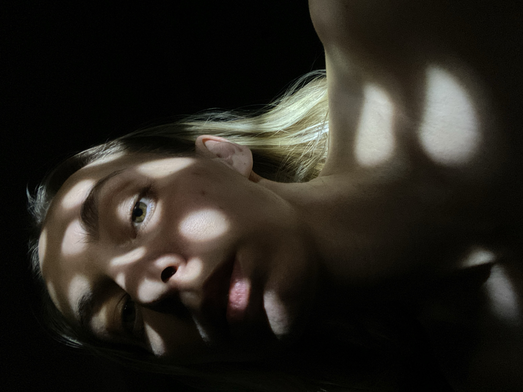 A close-up, low-angle shot of a light-skinned person's face and neck, with light blond hair visible near the ear and behind the neck. The person is in a predominantly dark environment, with roundish patches of light illuminating parts of the face and the side of the neck. The face is tilted, with the nose and mouth in the lower center of the frame. The person's left eye is visible and looking upwards. The image composition and content do not provide any indication of the location being the city of Chaikovsky, Russia.