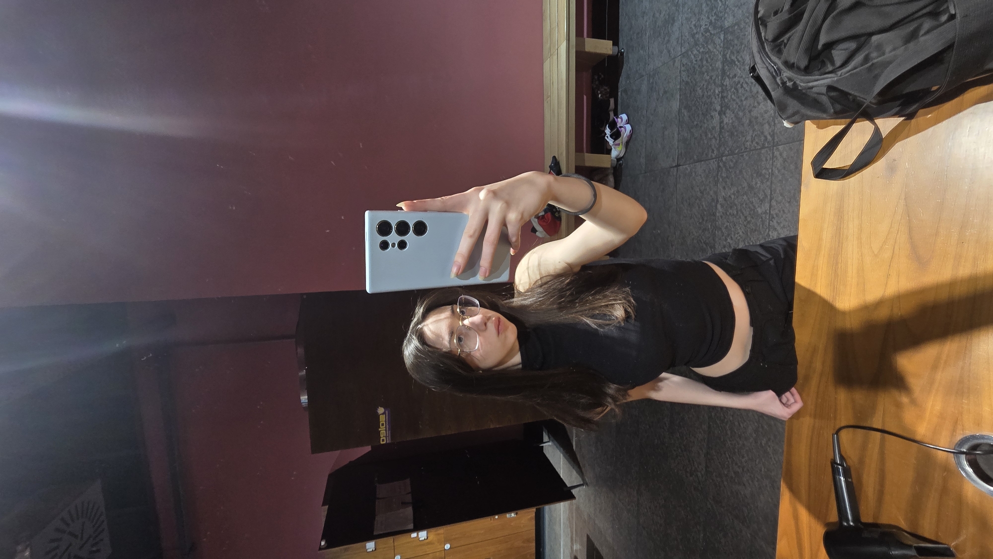 A young woman is taking a selfie in a mirrored surface that is reflective of what is behind her. She is wearing a black top and pants, and glasses, and holding a blue smartphone in her right hand. Her hair is long and brown. The mirrored surface reflects a red wall, some cabinets, and the logo of a business that reads "PSOAS". A wooden table with a black bag on it is visible to the right of the frame, and a hair dryer is on the table as well. Based on the presence of the PSOAS logo, it's likely this is inside a facility owned by the PSOAS organisation, which is active in Stara Zagora, Bulgaria.