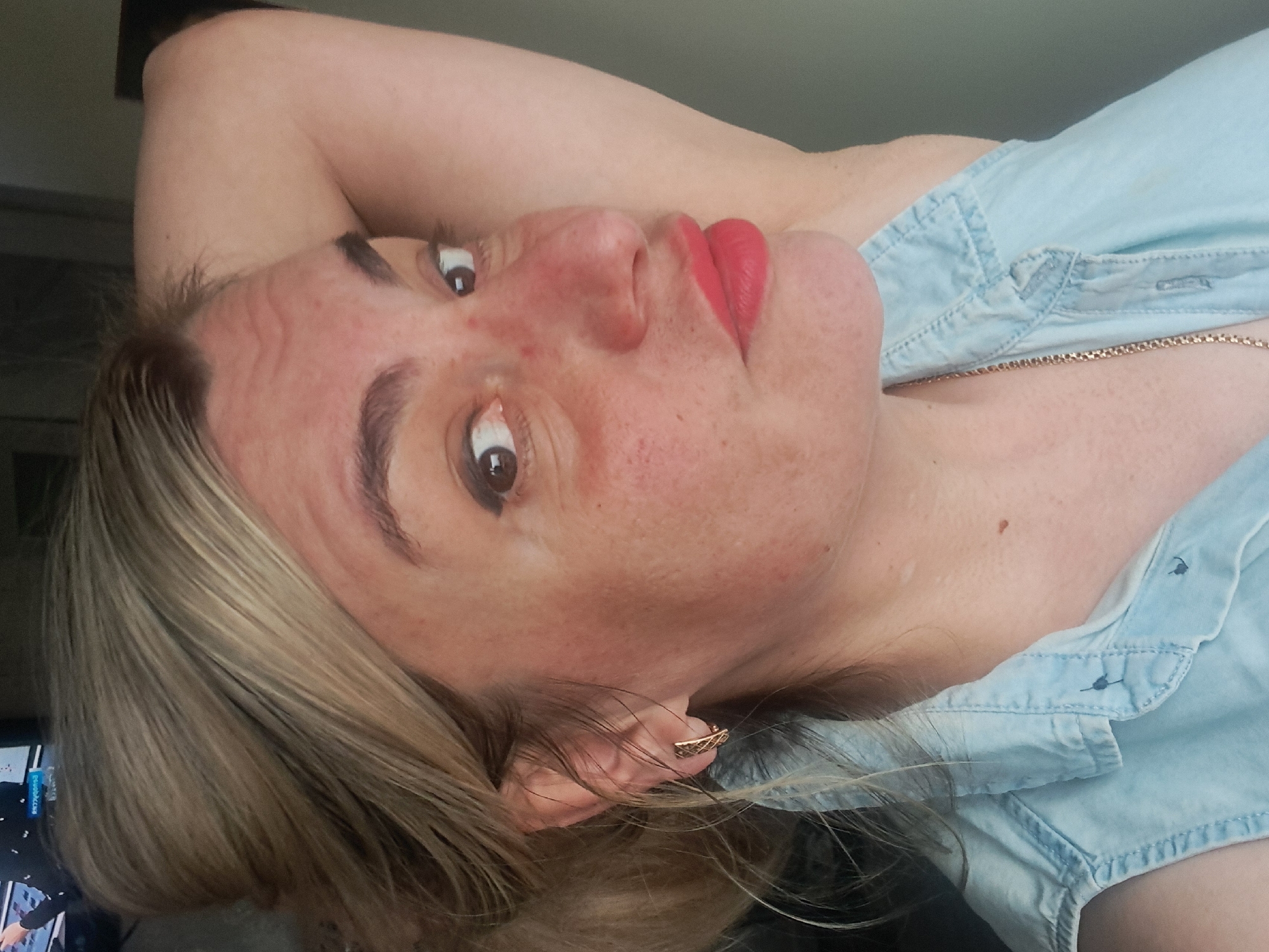 A woman is depicted in a close-up shot, her face turned towards the viewer. She has light brown hair with blonde highlights, and her eyes are brown. Her lips are painted a vibrant red. The skin of her face appears to have freckles or blemishes. She is wearing a light blue denim shirt and a delicate gold chain necklace is visible around her neck. An ornate gold earring adorns her ear. The background is out of focus, but a television screen with some writing and a person visible on it can be partially seen in the bottom left corner. There is no information about the location being in Elektrostal, Russia.