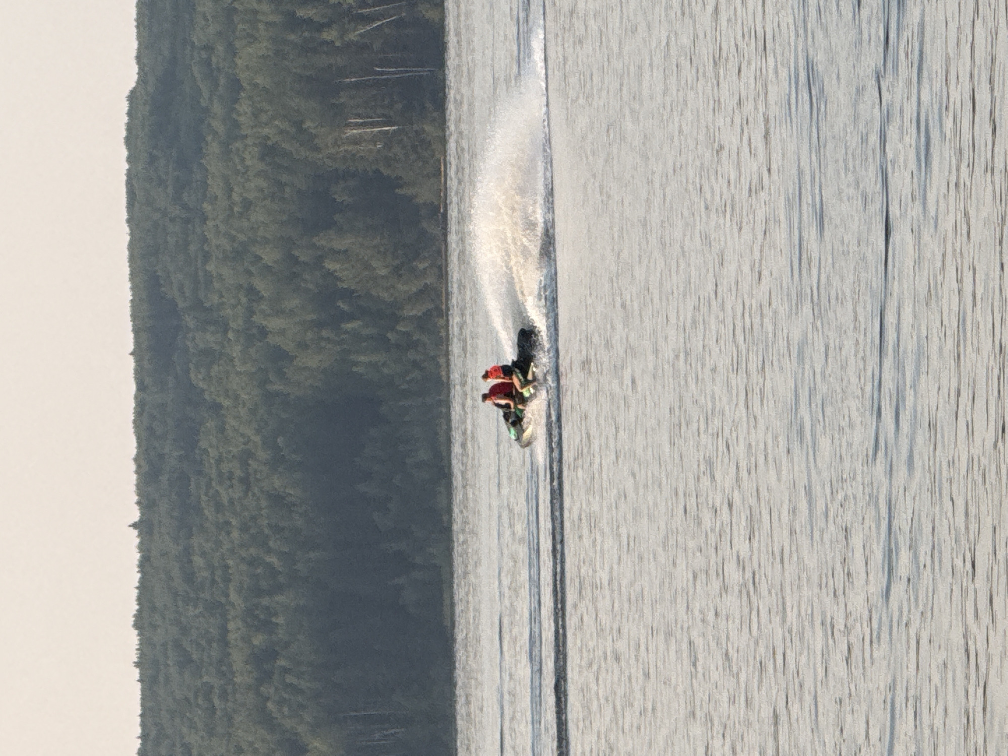 Two individuals, wearing red life vests, are riding a jet ski on a body of water. The jet ski is creating a large wake behind it. In the background, a dense forest covers a hill that slopes down towards the water. The sky above is a light, hazy color. This scene is likely taking place on the Kama River, near the city of Chaikovsky, Russia.