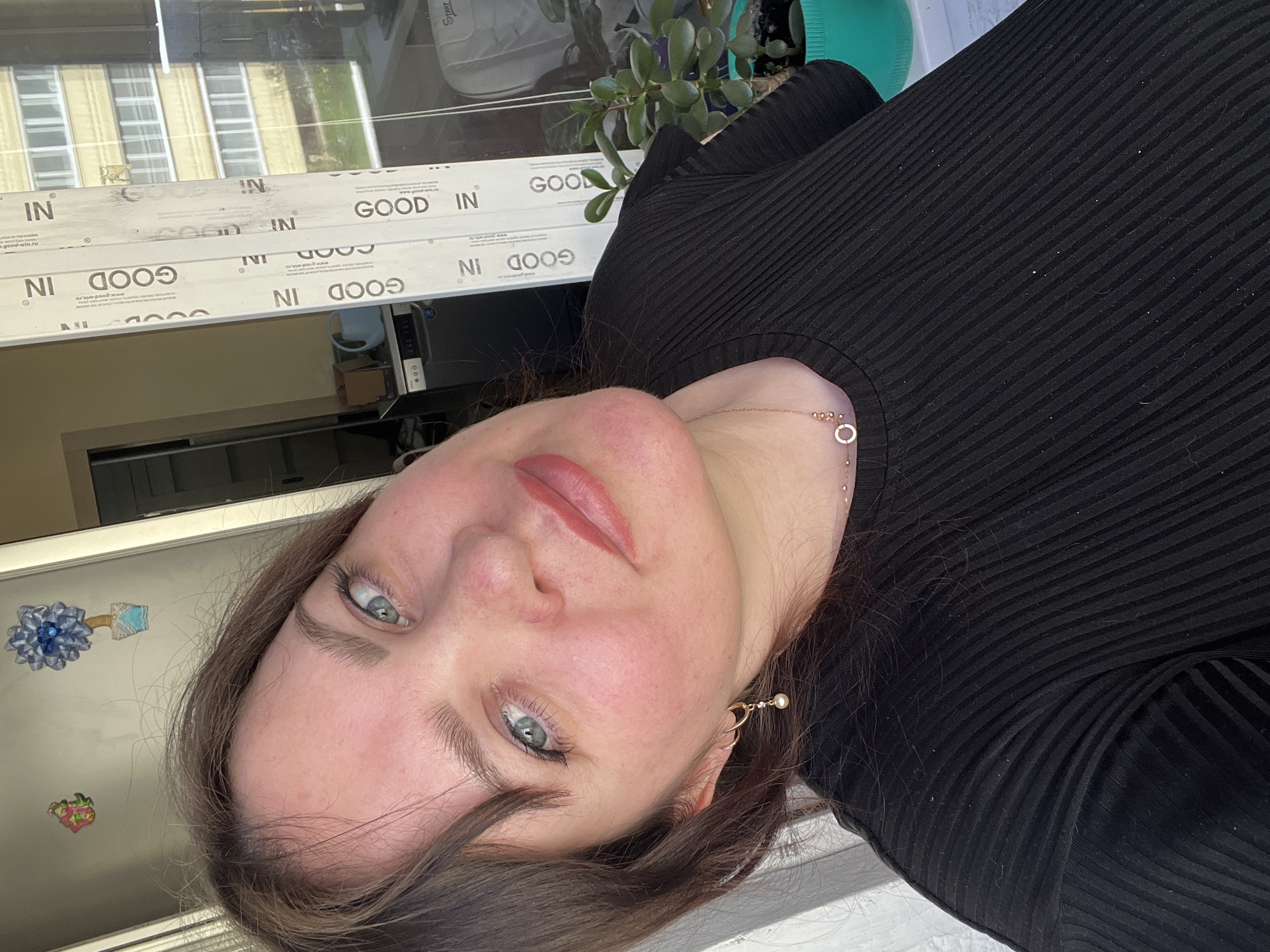 A woman is taking a selfie indoors. She has light skin, blue eyes, and brown hair. She is wearing a black ribbed top and gold jewelry. The background shows a window with the word "GOOD IN" repeated on the frame. Outside the window is a building. There is also a plant on the windowsill. The image does not provide explicit evidence of the location being in Noviy, Russia.