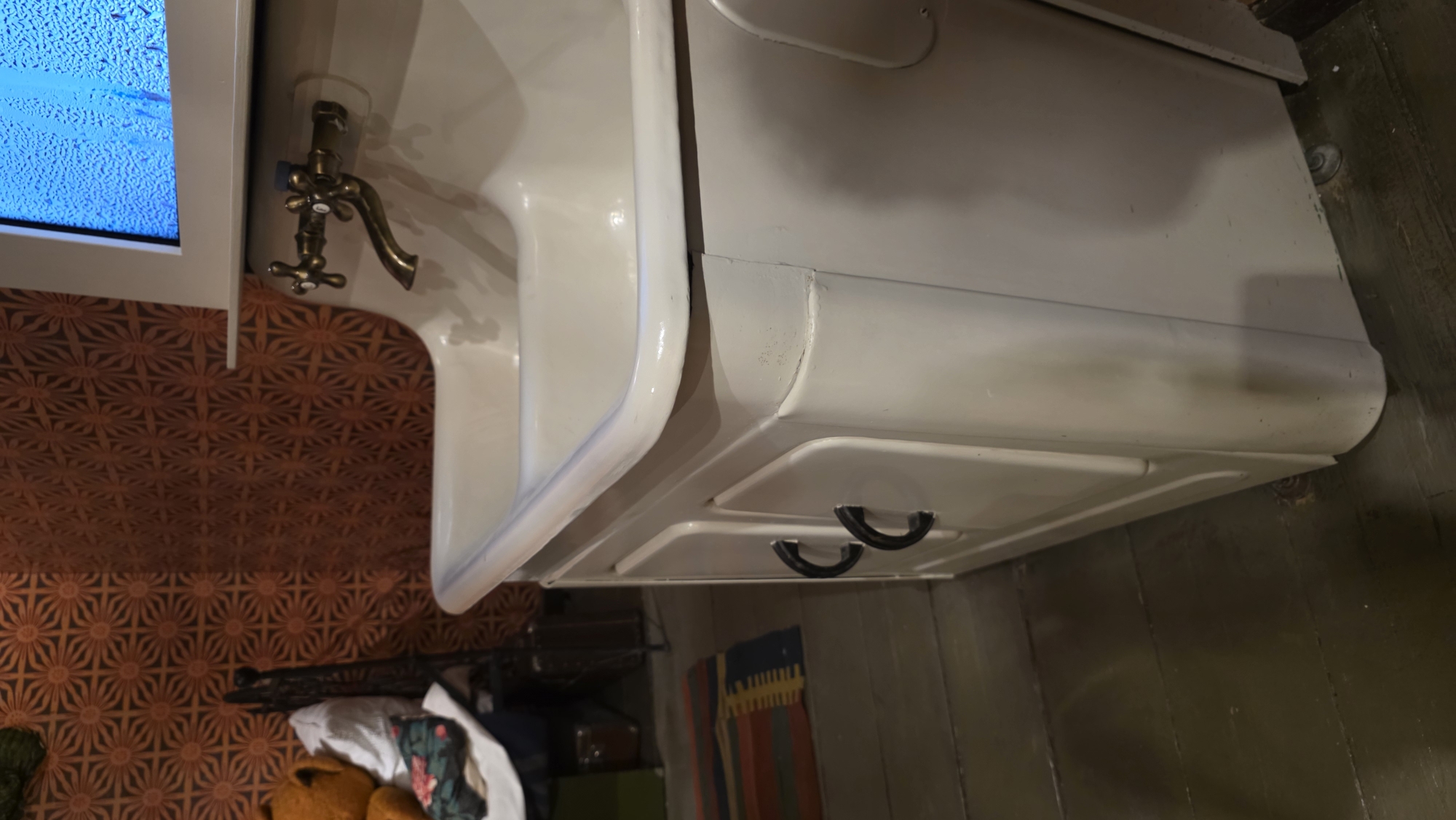 The image shows a white, vintage-style sink with a double-handle faucet, likely made of brass. The sink is part of a vanity with drawers, evident from the curved handles on one of the drawers. The backdrop consists of patterned wallpaper in shades of brown and orange. Below the vanity, a glimpse of the floor reveals wooden planks, along with assorted items like clothing, an object, and a striped rug. Without further information, location information is impossible to determine.