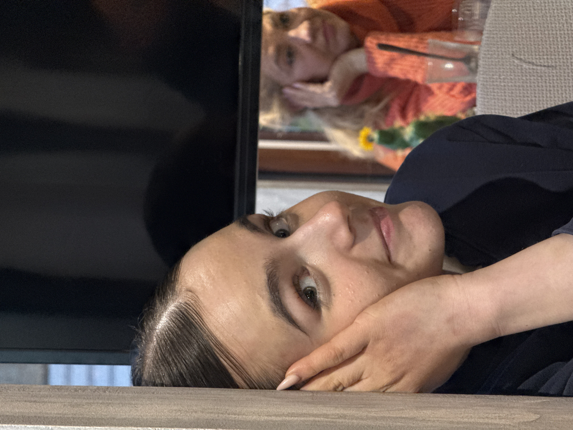 The image depicts an indoor scene in Sofia, Bulgaria, featuring two females. In the foreground, on the left, a young adult female is partially visible behind a light-colored vertical wooden or laminate panel. She has dark hair pulled back, a light complexion with visible facial pores, and green/hazel eyes looking towards the right. A slight smile is present on her lips. Her left hand, featuring light-colored, almond-shaped manicured nails, is placed against her left cheek and ear. She is wearing a dark blue, long-sleeved garment.
In the blurred background, seen through a window or reflection, an older adult female with blonde hair is visible. She is wearing an orange/red textured knit sweater, and her right hand rests on her jawline. On a table in front of her, a small yellow flower in a green vase or bottle is present, alongside a clear glass containing a dark stirring implement. A light-colored, possibly woven fabric, chair back is also visible. The environment is dimly lit, suggesting an indoor setting with ambient artificial lighting. The foreground subject appears to be looking off-camera to the right, while the background subject has a neutral expression.