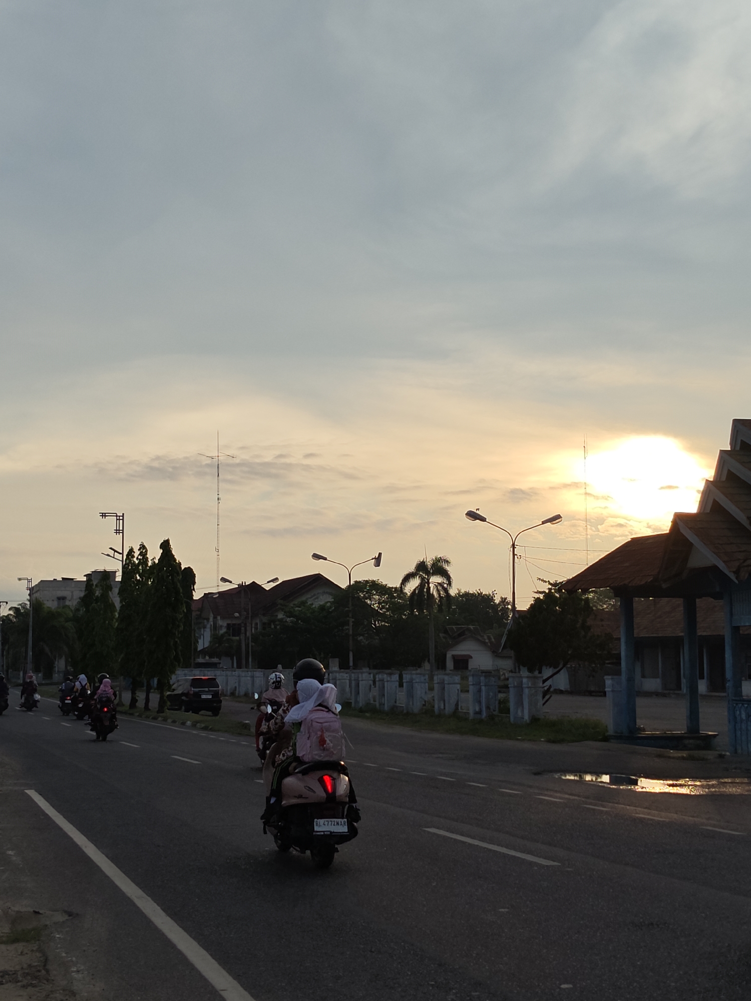 The image depicts a street scene in Lhokseumawe, Indonesia, under a bright, partially cloudy sky with the sun low on the horizon. Several motorbikes are traveling along the road, some carrying multiple passengers wearing head coverings and backpacks. Buildings and trees line the side of the road, along with streetlights. There is a bus stop with a roof structure and a puddle on the pavement reflecting sunlight. The light casts shadows on the road, creating an overall subdued lighting effect.