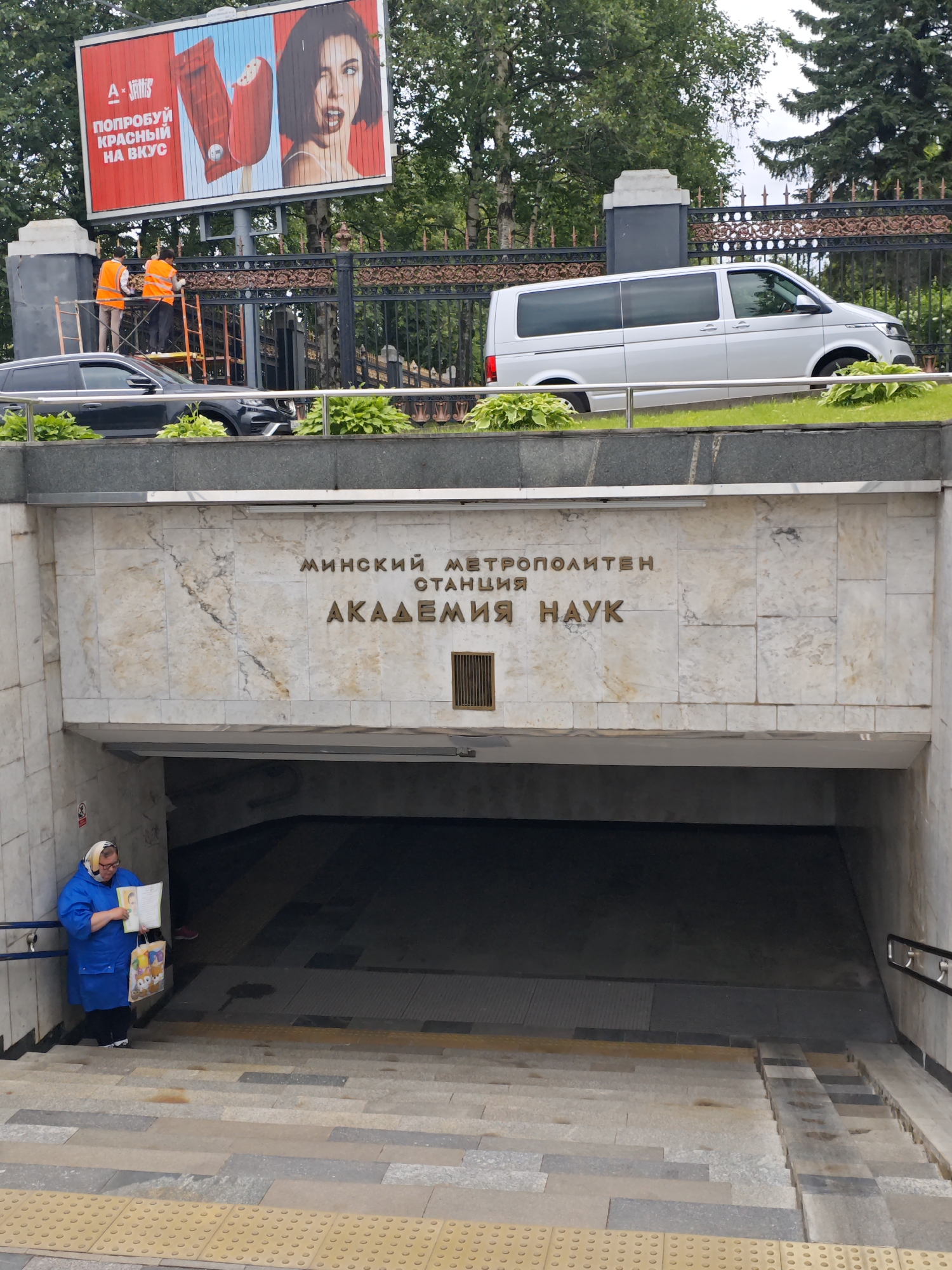 The image shows the entrance to the "Akademya Nauk" metro station in Minsk, Belarus. Above the entrance, a large billboard displays an advertisement for "A'Juniors" ice cream, featuring a close-up of two red ice cream bars and a woman with expressive eyes. Two construction workers in orange vests are visible on ladders near the billboard, appearing to be performing maintenance. A dark-colored car is parked below the billboard, and a silver minivan is visible further in the background.
The metro station entrance itself is framed by marble-clad walls with the station's name inscribed in metallic letters: "МИНСКИЙ МЕТРОПОЛИТЕН" (Minsk Metropolitan) and "СТАНЦИЯ АКАДЕМИЯ НАУК" (Station Academy of Sciences). Below the inscription, a metal grate is embedded in the wall.
On the left side of the stairs leading into the metro station, a woman wearing a blue coat and a headscarf is standing. She is holding a book open and appears to be reading, with two shopping bags at her feet. The stairs are made of stone and have yellow tactile paving at the bottom. The overall environment suggests an urban setting with a mix of commercial advertising, infrastructure, and daily life.
