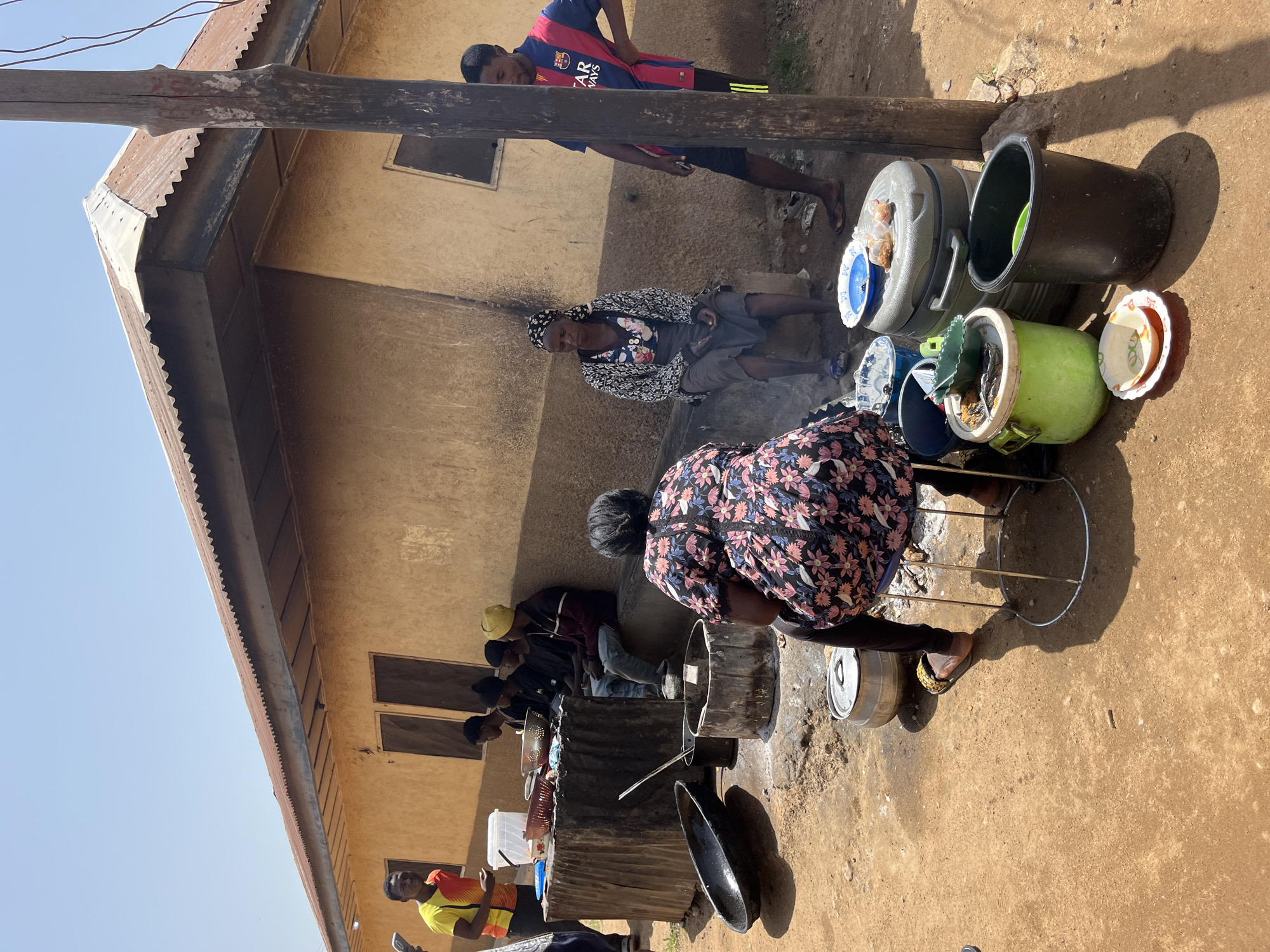 In Jos, Nigeria, a group of individuals are gathered outside a building with a corrugated metal roof. In the foreground, a woman in a floral dress sits on a metal stool facing away from the camera, tending to a cooking area. Around her are several cooking pots and containers, including a large black pot and a grey pot with a lid. To her left, a large, dark, oval pot rests on the ground.
Behind the woman, a row of young men and a woman are seated on a low concrete ledge. One man stands to the left, wearing a yellow and red shirt, appearing to be in conversation with a person off-camera. To his right, several young men are seated, some wearing black t-shirts and one in a yellow beanie. The woman next to them is wearing a patterned headscarf and a dark skirt. Further to the right, a young man in a blue and red soccer jersey stands near a wooden pole.
The environment is an unpaved, dusty area with a simple, rustic building as the backdrop. The building's walls appear to be made of earth or stucco, with some windows covered by wooden panels. Various plastic containers and plates are scattered around the cooking area, suggesting food preparation or serving. The scene conveys a sense of daily life and community activity in an outdoor setting.