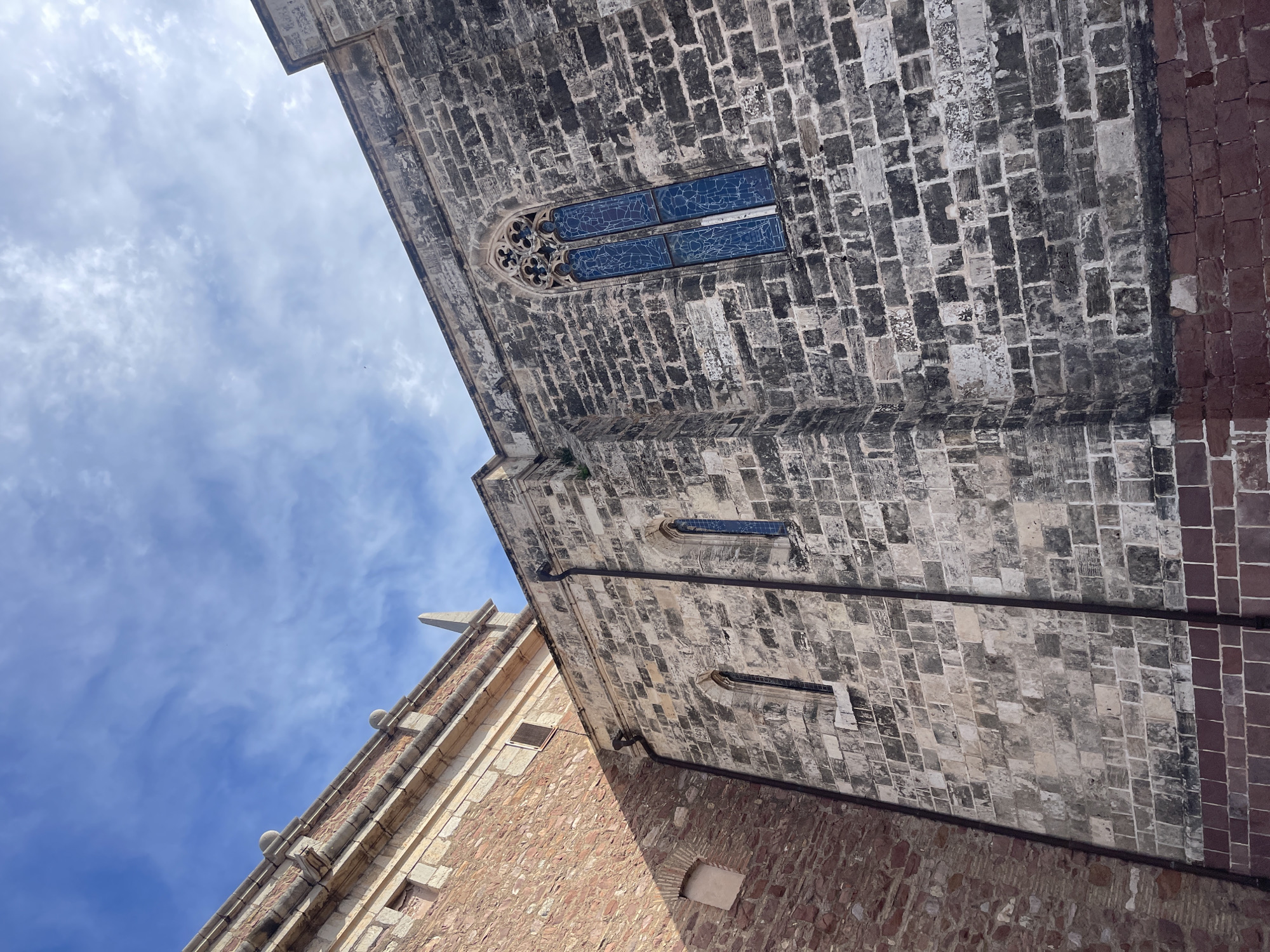 The image is a low-angle shot of a building in El Puig De Santa Maria, Spain. The building features a mix of grey and red brickwork. A large window with blue glass and ornate stone detailing is visible near the top of the frame. Below it, there are additional smaller windows. The sky is visible in the upper left corner, showing a blue sky with wispy clouds. The architecture suggests a historic or religious building, potentially part of a church or monastery. The overall perspective is upward, highlighting the building's height and architectural features.