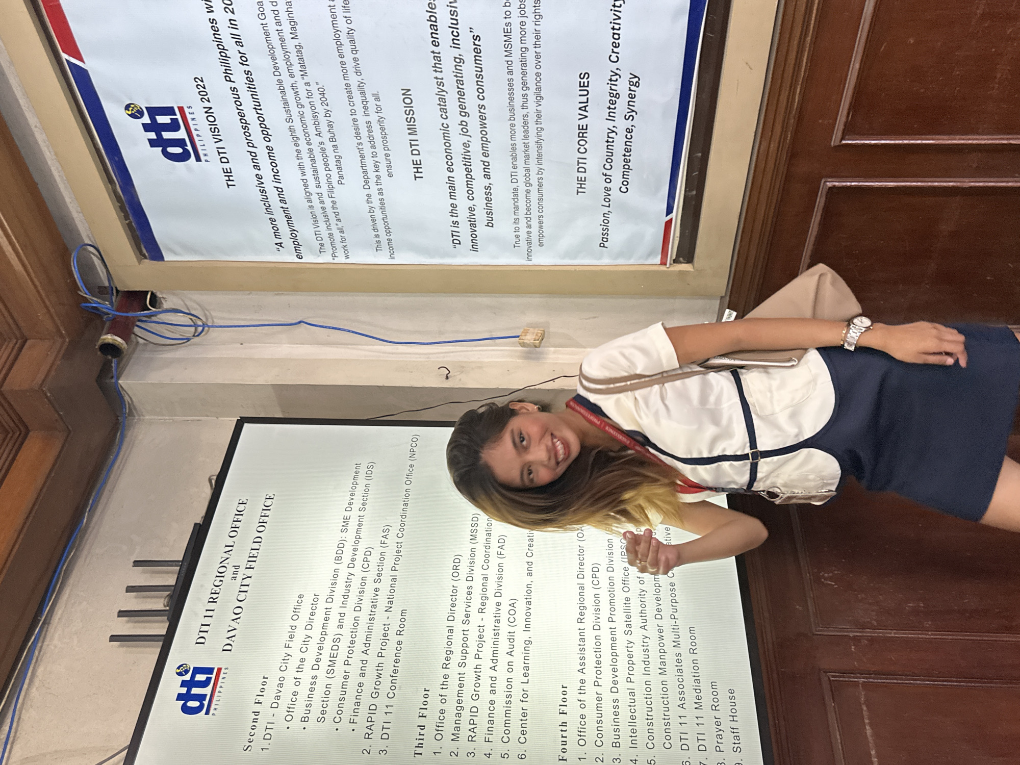 The image shows a young woman standing in front of a bulletin board with information about the DTI 11 Regional Office and Davao City Field Office. The bulletin board is divided into sections listing offices on the second, third, and fourth floors. There is also a section detailing the DTI Vision 2022, the DTI Mission, and the DTI Core Values. The woman is wearing a white blouse with a dark blue skirt and a lanyard around her neck. She is holding a beige bag on her shoulder and looking towards the camera with a smile. The environment appears to be an office hallway with wooden paneling on the lower part of the walls. The lighting is fluorescent and appears to be coming from overhead. The bulletin board has a matte finish and the text is in black. The DTI logo is visible in the upper left corner of the board.