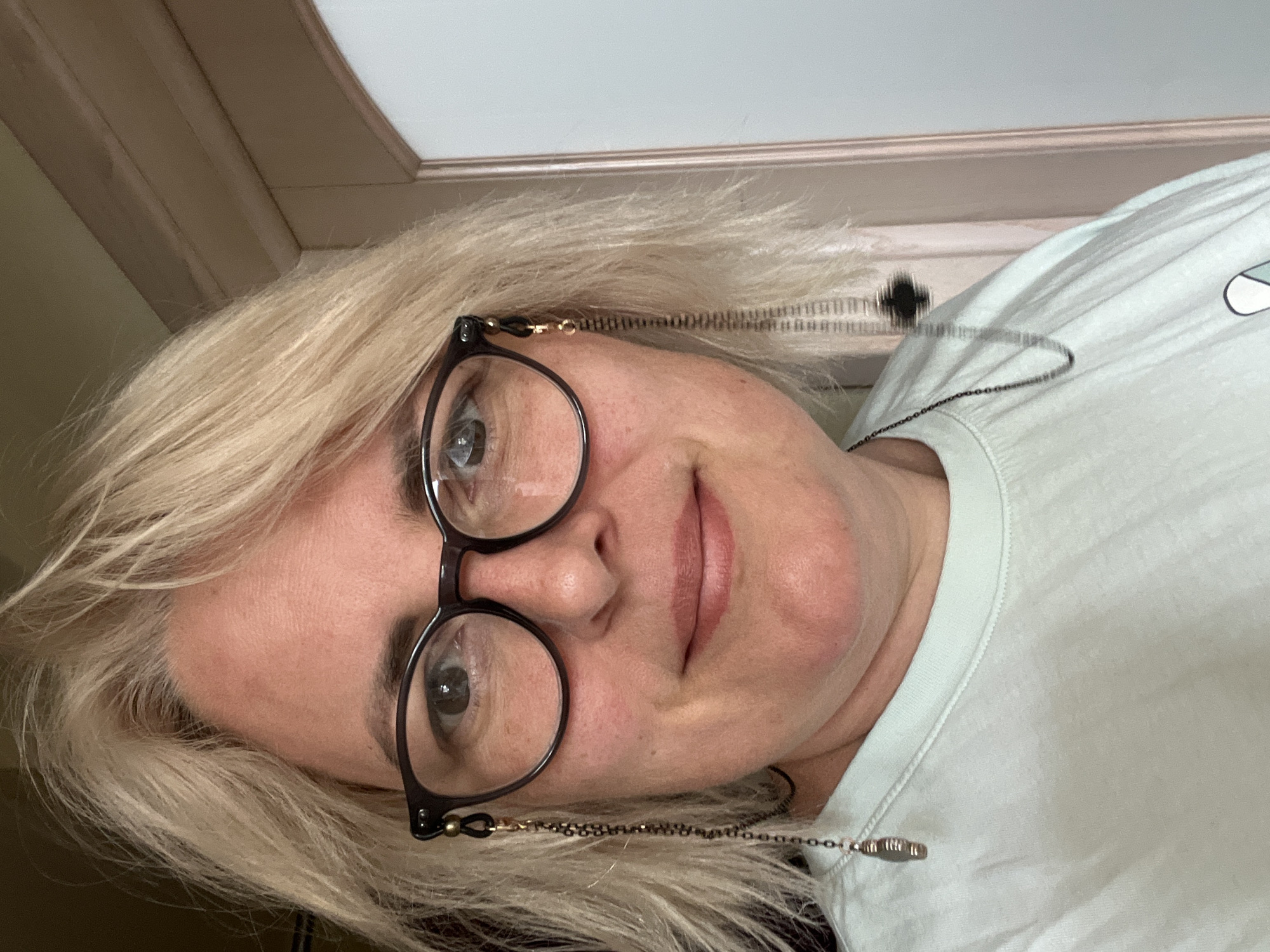 A light-skinned woman with blonde, choppy hair and glasses is looking at the camera. She is wearing a light blue t-shirt and her glasses have chains with charms. The background appears to be a doorway with a light-colored wall. The location is unknown.