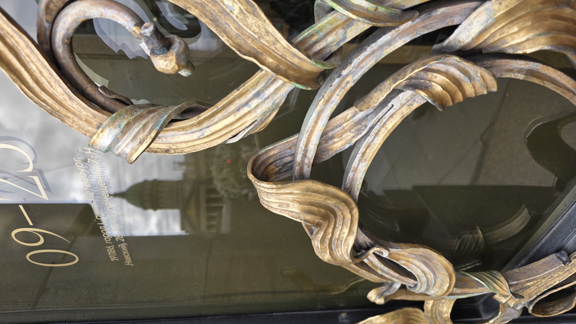 Close-up, low-angle, rotated view of a detailed, gold-colored metal architectural adornment on a dark, reflective glass surface with mirrored reflections of a building. In the lower left of the glass, there is partially visible gold lettering in Russian. The reflected building appears to have a green dome. The architectural element consists of curved, ribbon-like pieces of metal interwoven to form a decorative pattern. The metal shows signs of age and oxidation. The background is slightly out of focus due to the shallow depth of field. Based on the text in the glass, the location is St. Petersburg, Russia.