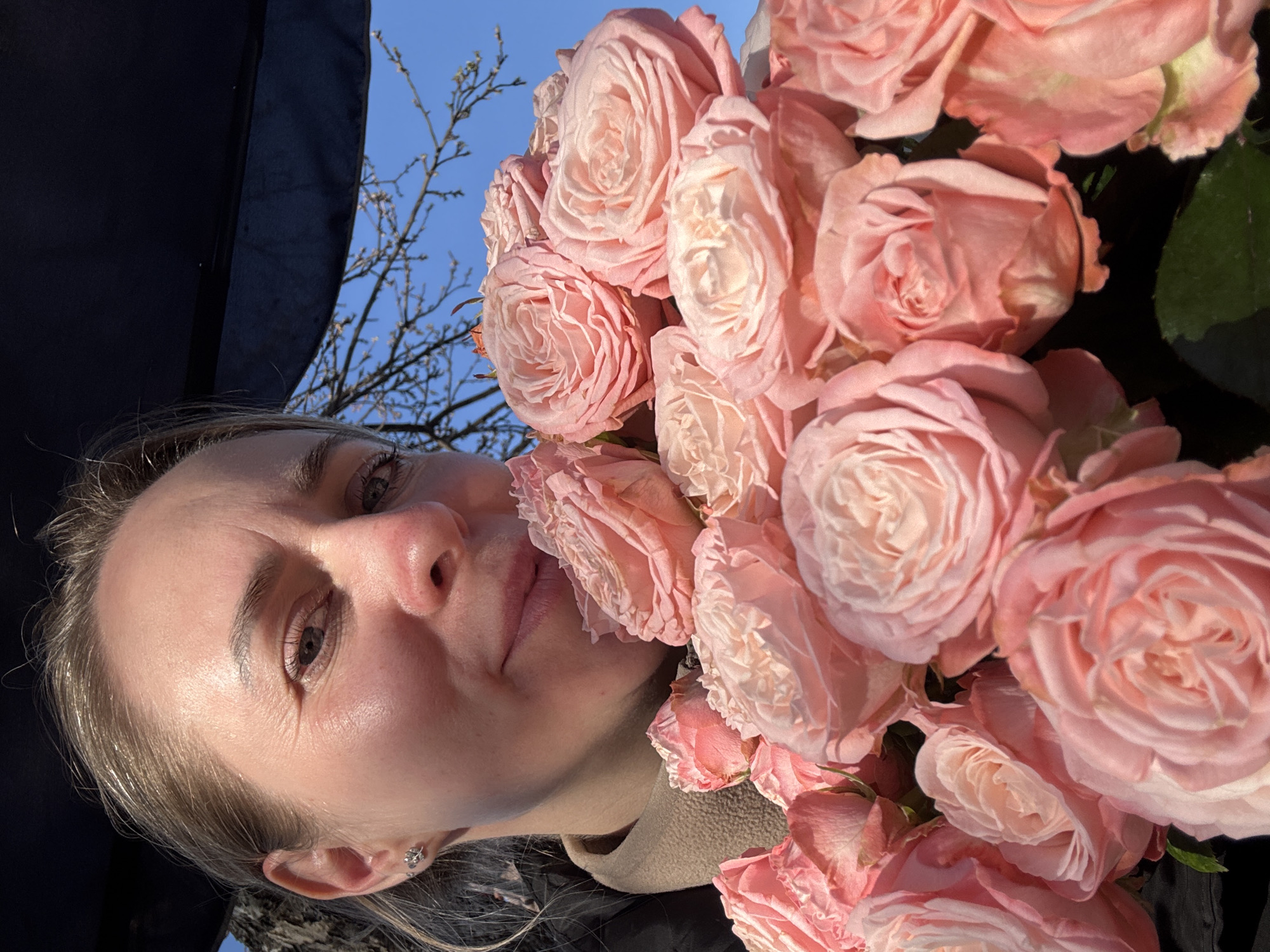 A light-skinned woman is holding a bouquet of pink roses up to her face. The background shows a clear blue sky and bare tree branches. The location is not discernible from the image.