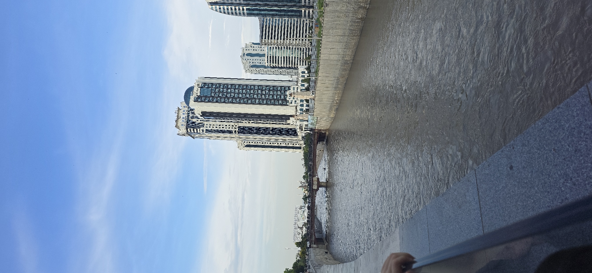 The image depicts a view of the Sunzha River in Grozny, Russia, with a modern cityscape in the background. The river occupies the lower half of the frame, its surface rippling with a murky, brown hue. The riverbanks are lined with concrete walls. A person's hand is visible on the left edge of the frame, resting on a granite embankment.
Dominating the right side of the background are several tall, contemporary buildings. The most prominent is a striking skyscraper with a black and white patterned facade and a rounded glass top. Adjacent to it are other high-rise structures, some with dark, reflective windows. A bridge spans the river in the mid-ground, with vehicles visible crossing it, suggesting it is a functional transportation link. Beyond the bridge, more buildings and the silhouette of minarets indicate the presence of more distant urban architecture. The sky above is a pale blue with scattered, wispy clouds. The overall scene conveys a sense of urban development alongside a natural waterway.