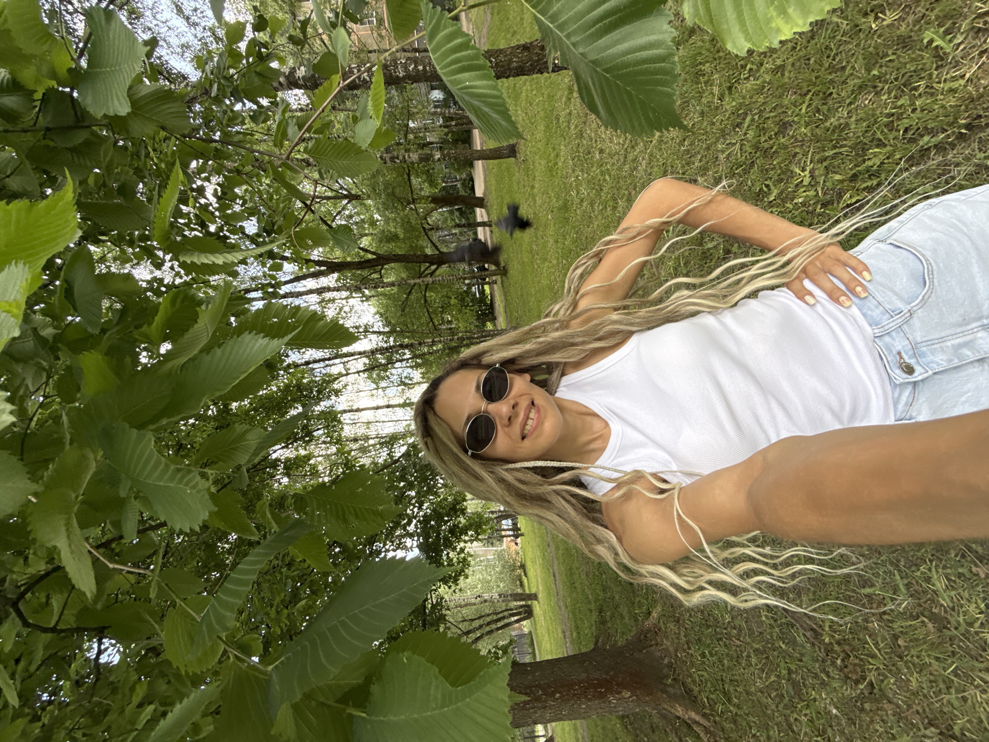 An outdoor, eye-level, full-body selfie is captured from the perspective of a young woman with long, blonde, curly hair, wearing oval sunglasses, a white tank top, and light-wash denim shorts. She is standing in a park-like setting with green grass, mature trees, and some foliage in the foreground and background. Sunlight filters through the leaves, creating dappled lighting. Two birds are in mid-flight in the background. The woman has her left hand on her hip and her right arm extended to hold the camera. The location is identified as Rusanovo, Russia.
