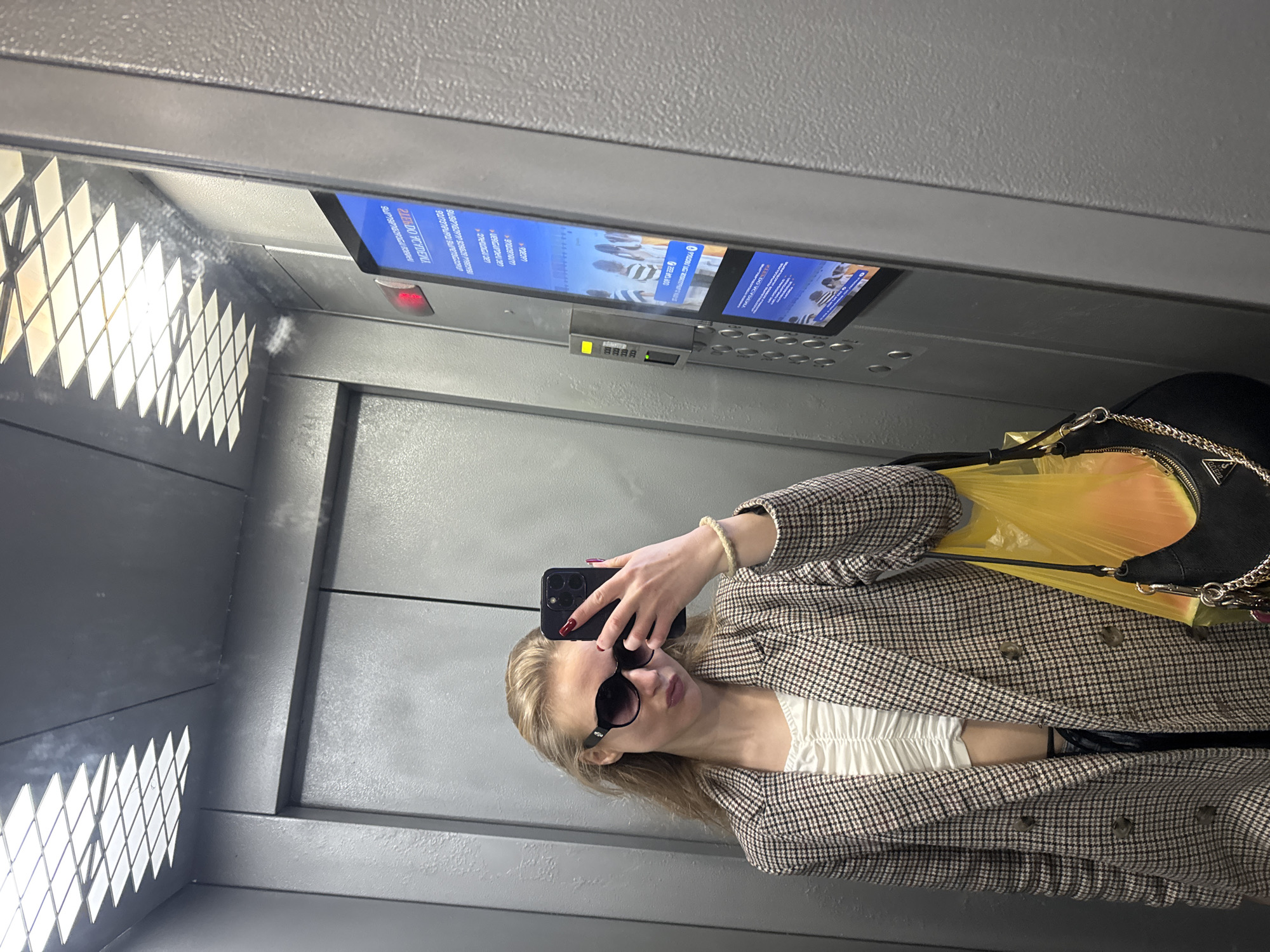 A woman is taking a selfie in an elevator. She is wearing sunglasses and a plaid blazer over a white top. A black purse with a gold chain is hanging on her shoulder. There is a yellow plastic bag next to the purse. Above her head is a digital display showing an advertisement. The elevator interior is gray with geometric patterns visible on the left. The location is Unknown, Unknown.