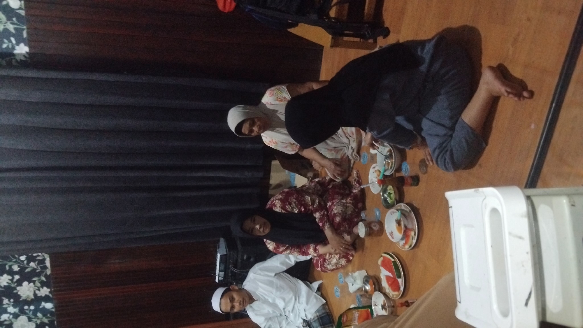The image depicts an indoor scene with four individuals seated on a wooden floor, arranged around a low table laden with food. The environment appears to be a modest dwelling in Bangun Rejo, Indonesia.
On the left, a man dressed in a white koko shirt and a peci (a traditional cap) sits facing left. Next to him, a woman in a maroon floral pattern dress and a black hijab is seated, looking towards the camera. To her right, another woman in a light-colored patterned blouse and a grey hijab is seated, looking down. In the foreground on the right, a person wearing a black hijab and a grey outfit is seated on the floor, with their back partially to the camera and their legs extended.
The table holds several plates of food, including sliced watermelon, bowls of what appears to be soup or stew, and some smaller dishes. There are also bottles and glasses on the table. A white cooler or storage container is visible in the lower left portion of the frame. The background features dark grey curtains, wooden paneling, and a floral patterned wallpaper at the top left. The lighting is dim, suggesting an evening setting. The overall atmosphere is one of a casual gathering or meal.
