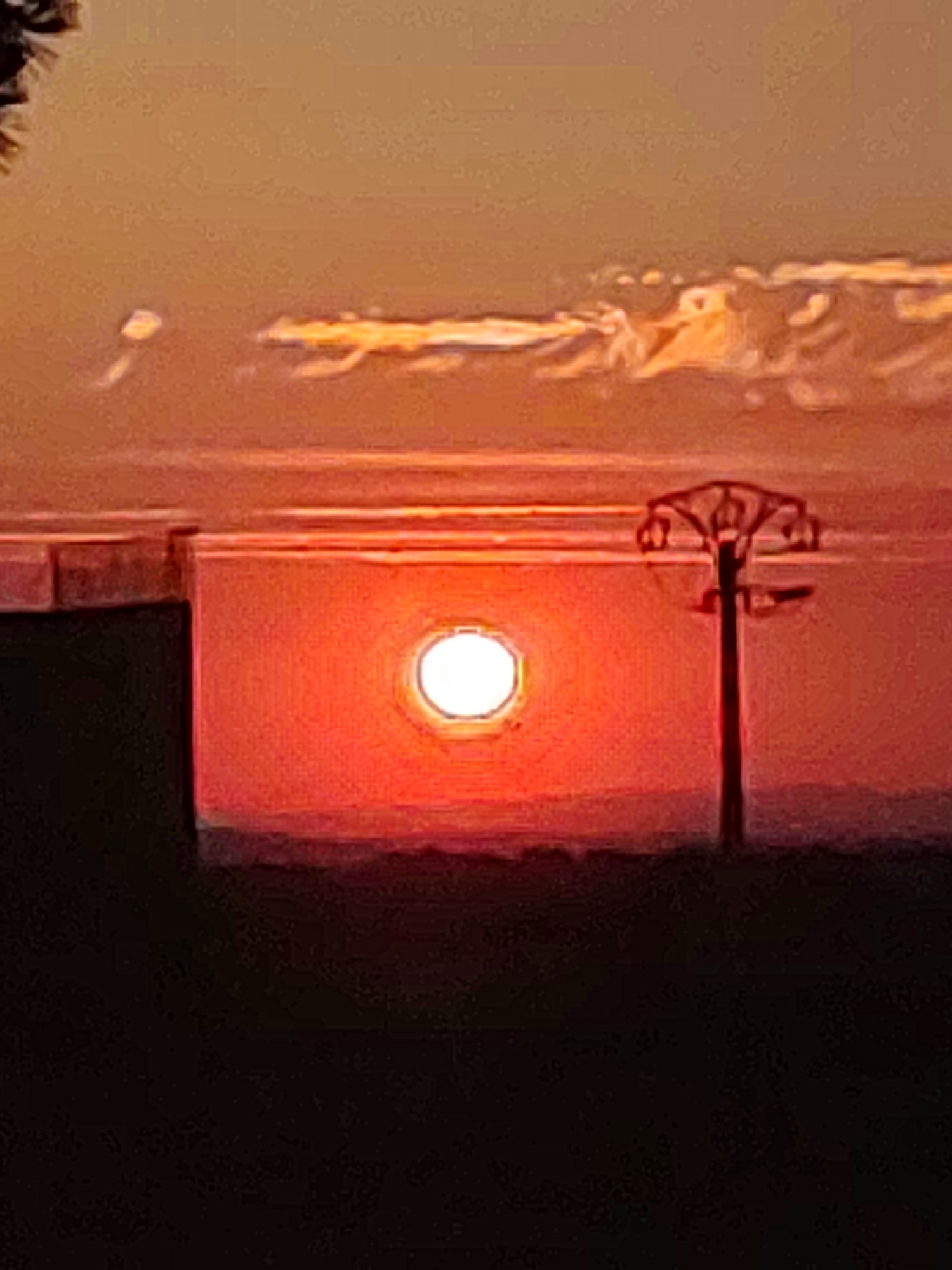 This image captures a sunset over the town of Sobradillo, Spain. The sun, a bright orb with a distinct oval shape, is low in the sky, casting a warm, reddish-orange glow across the landscape. In the foreground, a silhouette of a power line tower stands against the vibrant sky. To the left, a dark, rectangular structure, possibly a building or wall, is partially visible. The horizon is defined by a faint, darker line, suggesting distant hills or terrain. The sky itself is a gradient of warm colors, with wispy clouds illuminated by the setting sun. The overall impression is one of a peaceful, rural evening.