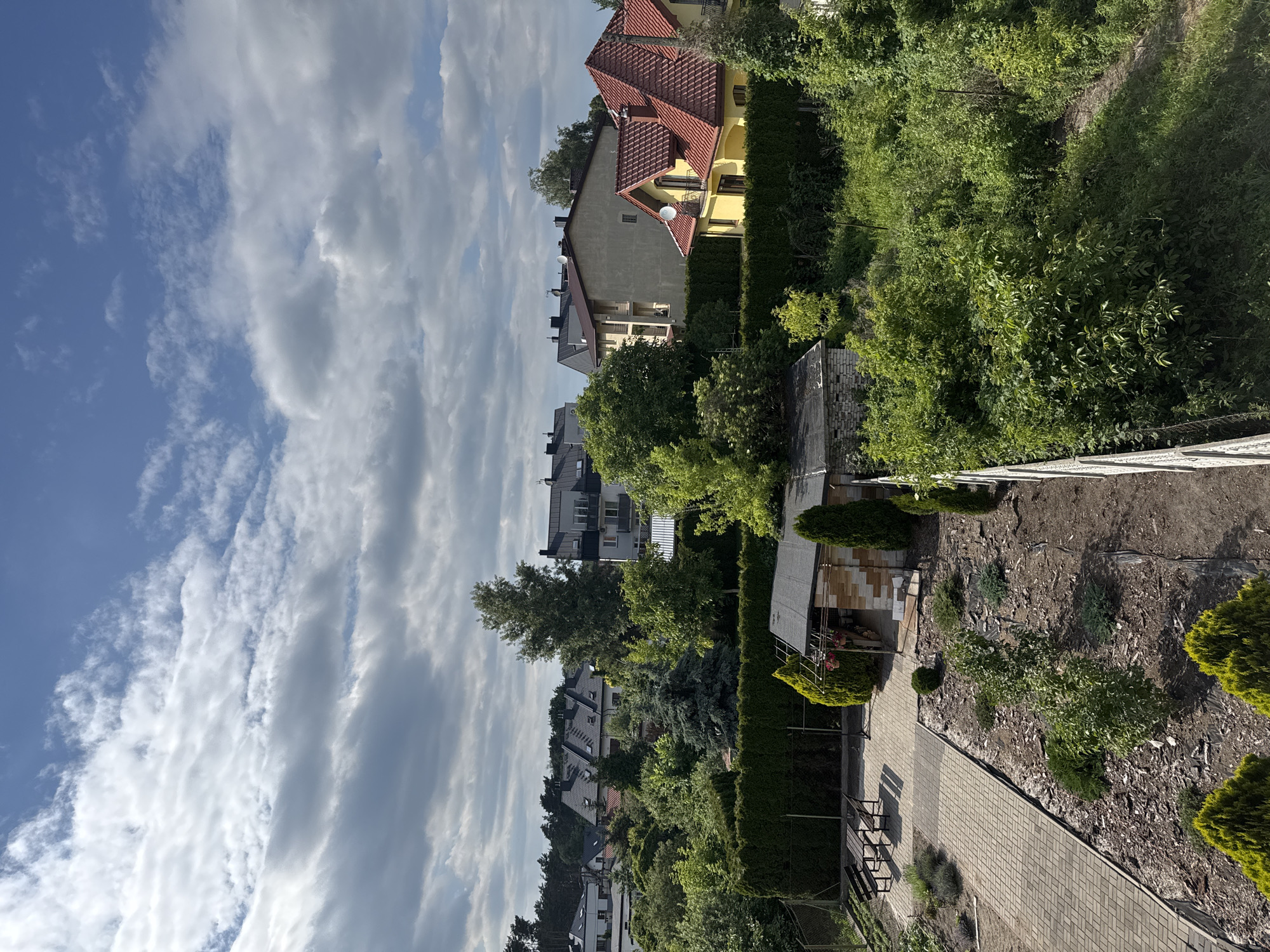 This image depicts a suburban backyard scene in Marki, Poland. The setting is a residential area with multiple houses and lush greenery. The foreground features a paved walkway and a garden area with mulch, small shrubs, and some larger manicured bushes. A small, rustic shed or outbuilding is visible in the middle ground.
Beyond the immediate garden, several houses are partially visible. One house, painted yellow with a red tile roof, is prominent in the upper portion of the frame. Other houses with various rooflines and architectural styles are seen further back, interspersed with dense trees and hedges, suggesting a well-established neighborhood.
The sky above is a mix of blue and white clouds, indicating daytime with partly cloudy weather. The sunlight appears to be casting shadows, suggesting it could be late afternoon or early morning. There is no activity or event taking place; it's a static view of a backyard. No people are visible, and no text is present in the image. The overall impression is one of a peaceful, green, and well-maintained residential environment.