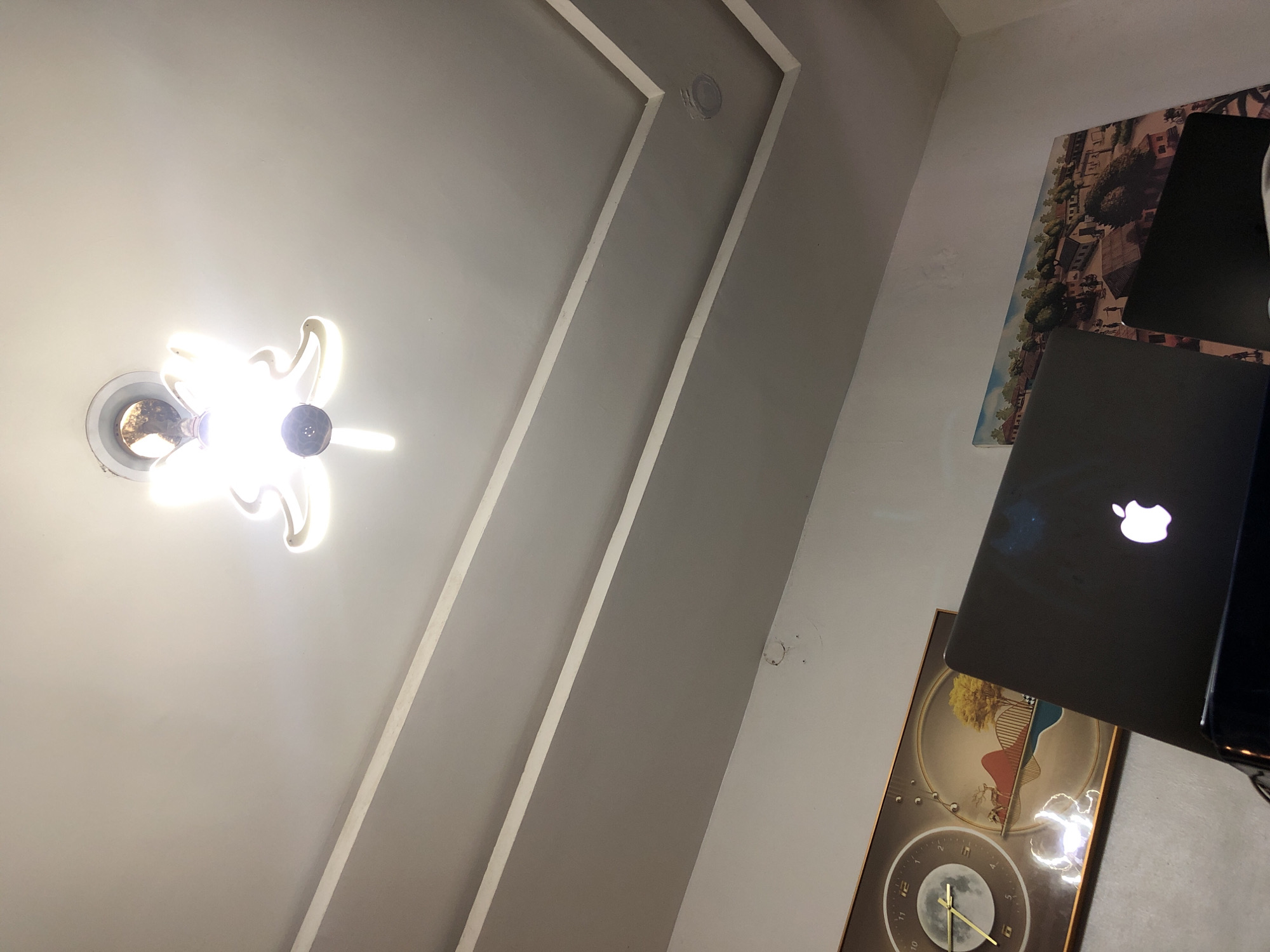 The image presents an interior view, primarily focusing on a ceiling and an adjacent wall, captured from a low, upward-angled perspective. A prominent, bright ceiling light fixture, possibly a multi-bulb chandelier with a distinctive ornamental design, emanates intense white light from the upper-left area. The ceiling itself features a contemporary false ceiling design, characterized by white linear moldings contrasting against a light gray background. A small, circular recessed element, possibly a speaker or sensor, is visible within the gray ceiling panel.
On the right side of the frame, a dark gray MacBook laptop is open, its white Apple logo brightly illuminated, suggesting it is powered on. Below the laptop, a large rectangular wall hanging, which appears to be a decorative clock or an art piece, is partially visible. It features a golden tree motif at the top and a circular moon-phase dial with Roman numerals and golden hands at the bottom. Above the laptop, further up on the wall, is a framed picture or painting depicting a detailed, elevated view of a town or village with buildings and lush trees. The strong artificial lighting from the ceiling fixture implies an indoor setting, likely during the evening or night. No individuals are present in the scene.