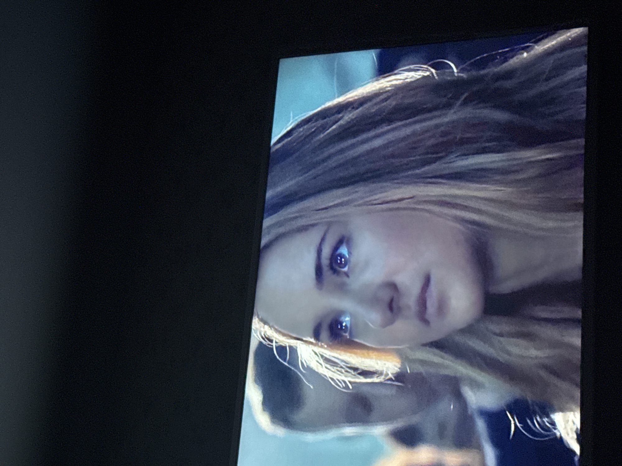 The image shows a close-up of a woman's face on a screen, possibly a television or monitor. The woman has light skin, blonde hair, and blue eyes. Her expression appears serious. Part of another person's face is visible in the lower portion of the screen. The background appears to be a light blue color. The image is framed by the dark border of the screen itself. The location of Mytishchi, Russia is not relevant to the visible content of the image.