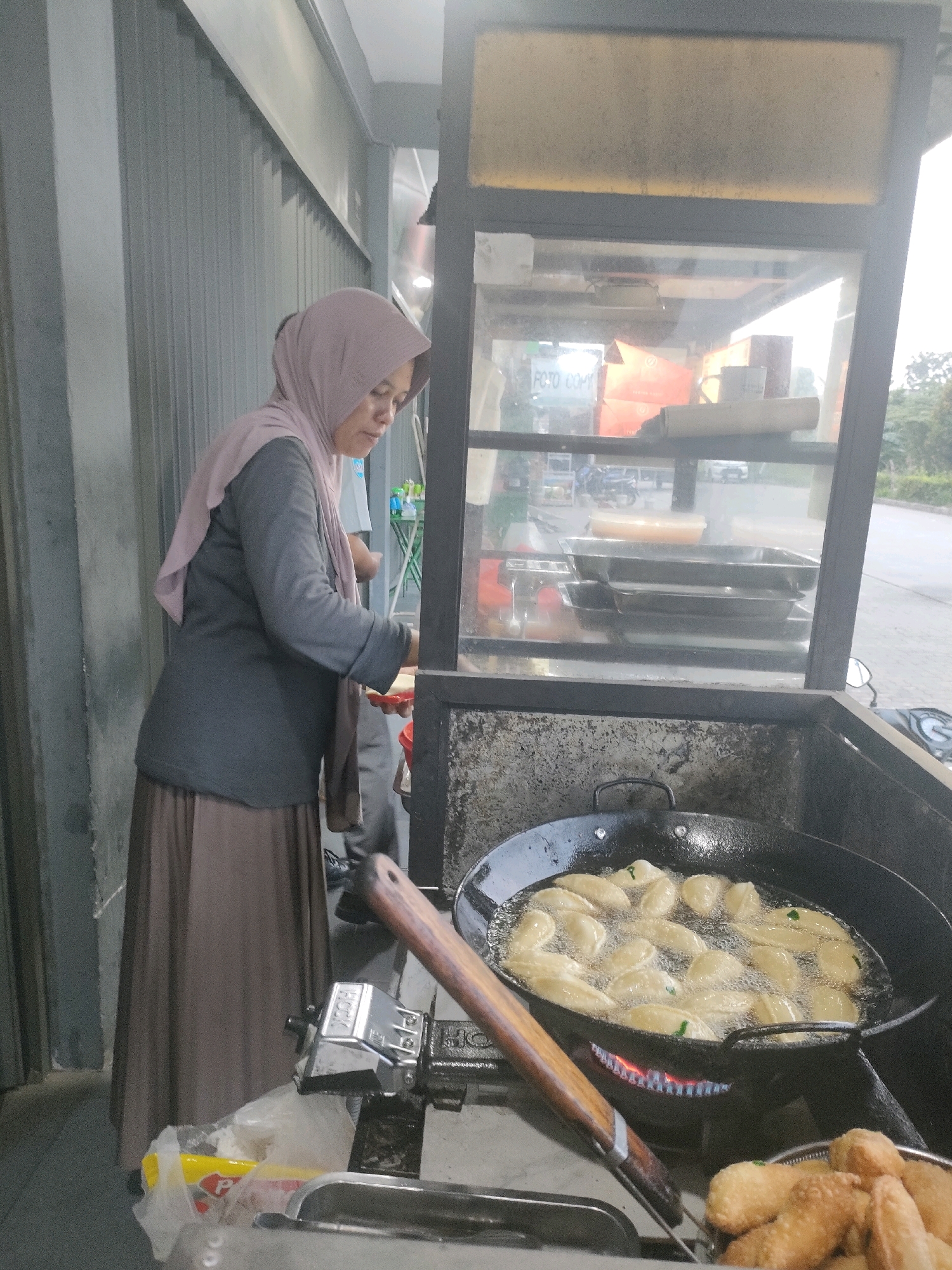 The image depicts an outdoor food stall likely situated in Batam, Indonesia, given the presence of common street food preparation. A woman wearing a light pink hijab and a dark grey long-sleeved shirt over a brown skirt is standing next to a food stall display. She is looking downwards and appears to be in the process of preparing food. In the foreground, a large black wok filled with numerous fried dumplings or similar items is placed on a gas stove with visible flames. A wooden-handled spatula is resting on the edge of the wok. To the right of the wok, a metal basket contains fried food items. The food stall has a glass display case above, showing stacked metal trays and some boxed items inside. The background reveals a street scene with trees and vehicles, suggesting an urban setting. A grey corrugated metal wall is visible on the left side of the image. The overall impression is of a busy street food vendor at work.