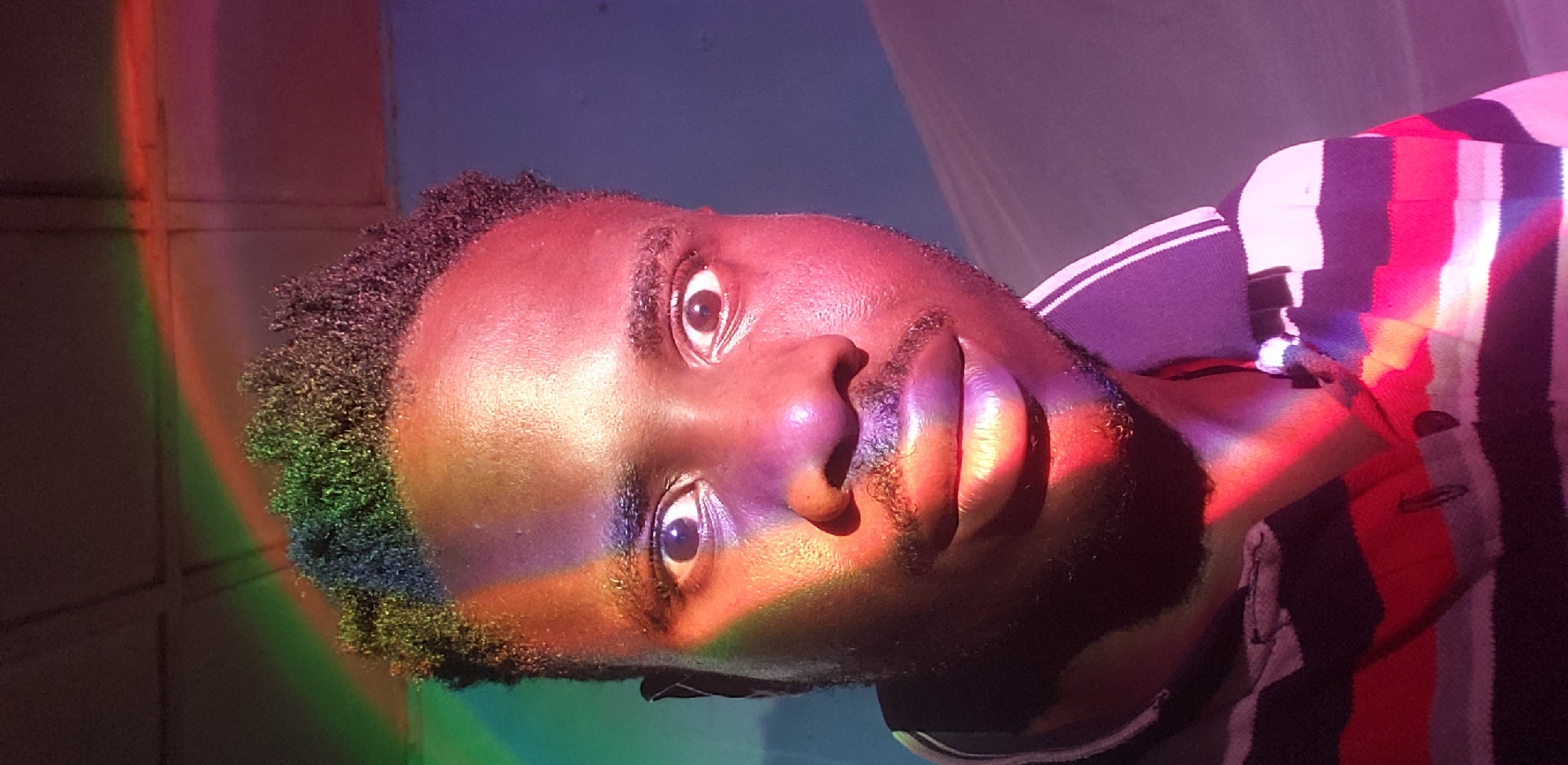 In Narayi, Nigeria, a close-up, front-facing selfie captures a young man's face illuminated by a spectrum of colored lights, creating a rainbow-like effect across his skin and hair. He has dark, curly hair and dark eyes, which are wide open and looking directly at the camera. A light mustache and goatee are visible. He is wearing a collared shirt with horizontal stripes in black, white, red, and possibly other colors. The background appears to be a plain wall, with a hint of a ceiling structure at the top. The lighting is the most striking element, casting vibrant hues of red, green, blue, and purple onto the subject and his clothing.