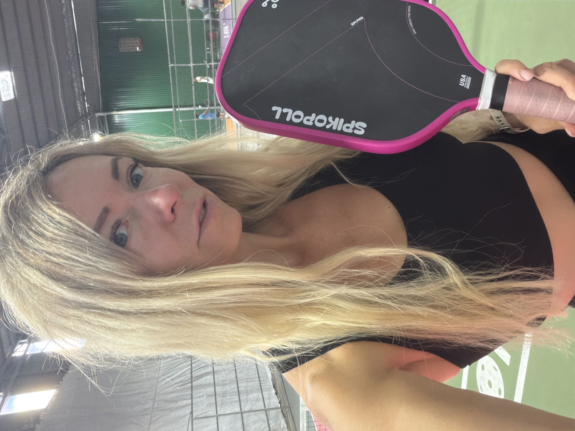 A woman with blonde hair, wearing a black tank top, holds a pink and black pickleball paddle. She is standing on a green pickleball court. The background shows a building with a metal roof and green walls, with other people playing pickleball in the distance. There is a white line and a circular marking on the court. The scene appears to be indoors. The specific location in Da Lat, Vietnam is not discernable from the image.