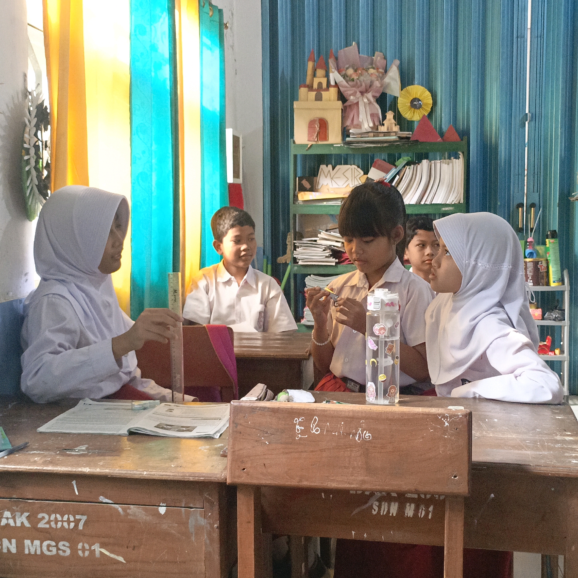 The image depicts a classroom in Salatiga, Indonesia, likely an elementary school, with four students visible. On the left, a female student wearing a white hijab and uniform is looking down at a ruler she holds in her right hand, positioned over an open book on her wooden desk. To her right, a male student in a white shirt uniform looks towards the front of the room. In the center right, another female student, also in a white hijab and uniform, is focused on manipulating small objects in her hands, with a clear plastic water bottle adorned with stickers on her desk. Behind her, a male student's face is partially visible. The classroom environment is characterized by wooden desks, a bookshelf filled with books and decorative items including a castle model and a paper flower, and colorful curtains in the background that allow light to enter the room. The desks have visible markings and labels, such as "AK 2007" and "N MGS 01," suggesting the school's identity or a designation related to the academic year and school name. The overall scene conveys a sense of a typical school day with students engaged in learning activities.