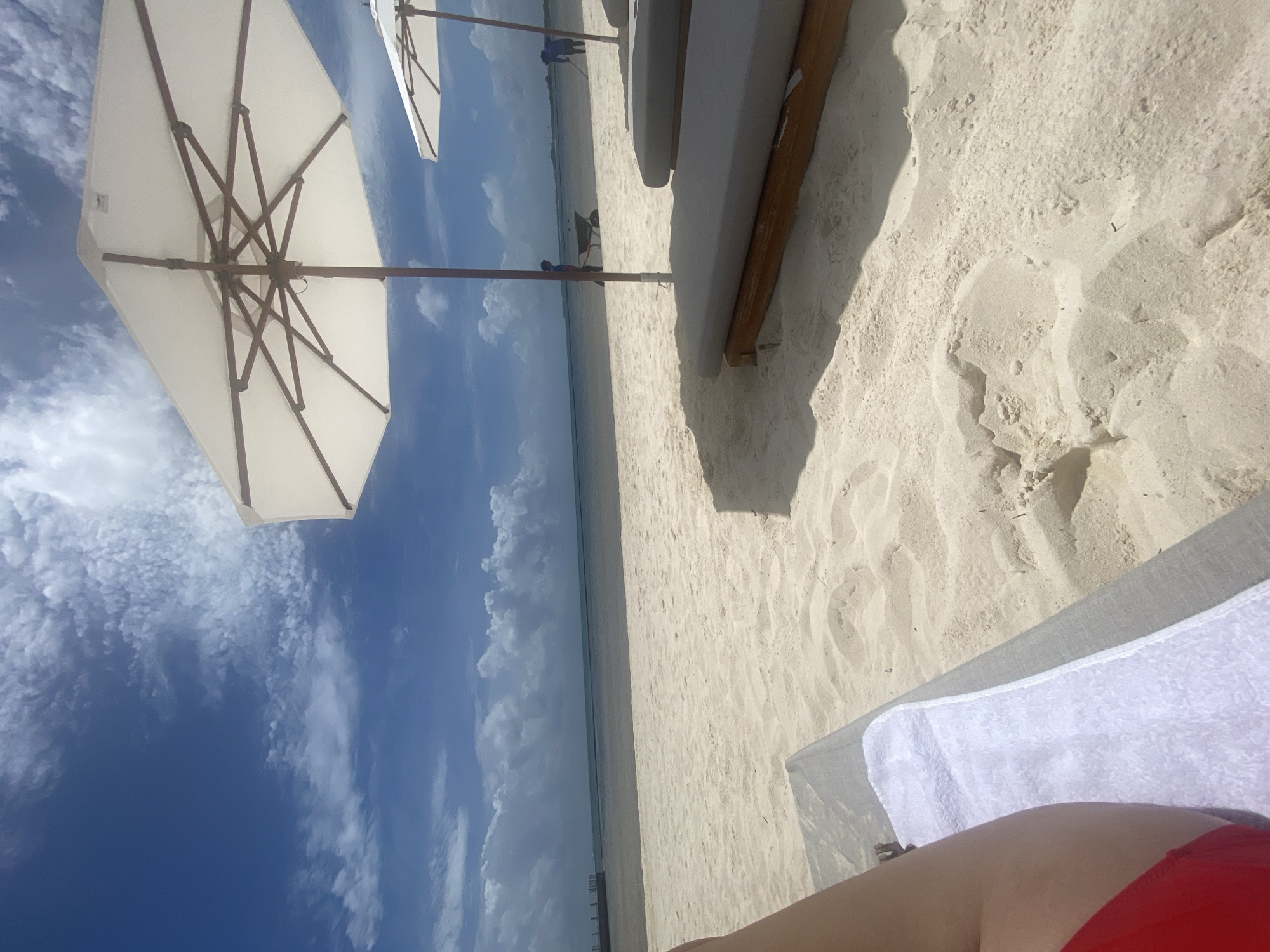 View from a lounge chair at the beach in Marina Maroma, Mexico. A white umbrella is visible to the left, and blue sky and clouds are in the background. The beach is white sand with some lounge chairs on it. The ocean water is visible in the distance. The legs and red bathing suit of the person lying in the lounge chair are visible in the foreground.