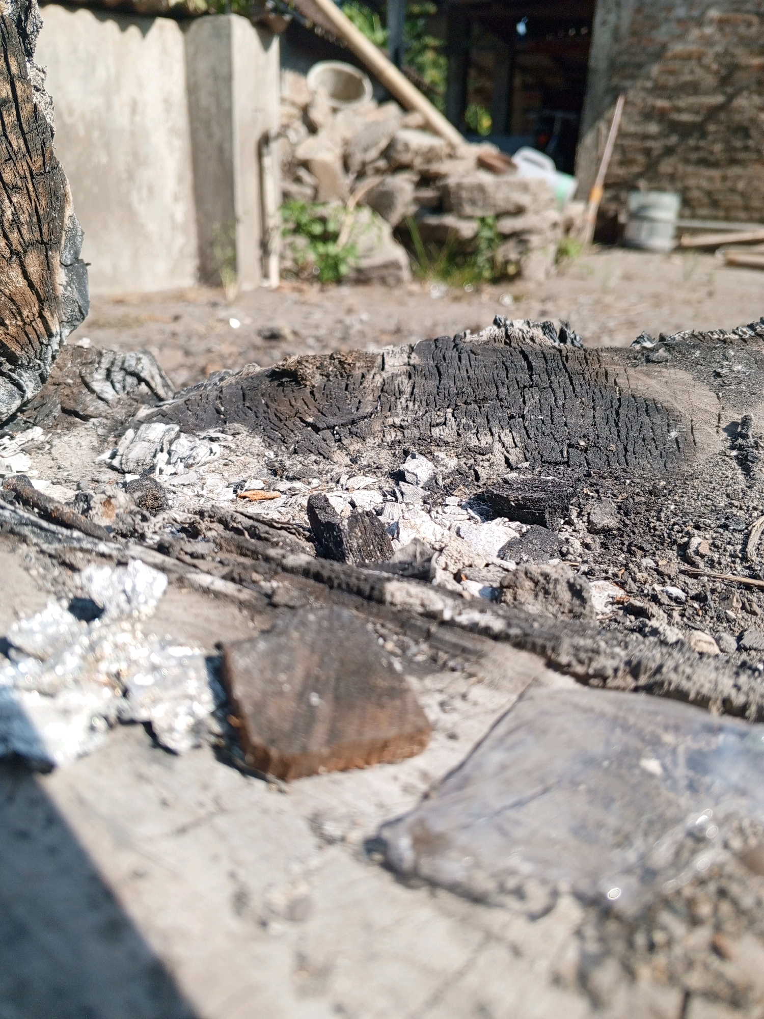 The image shows a close-up of burnt wood and ash on a wooden surface, with a blurred background depicting an outdoor scene. The background includes a concrete structure, a pile of stones, greenery, and a building with a brick facade. The lighting suggests it is daytime. While the image is set in Bantul, Indonesia, there are no specific landmarks or contextual clues within the frame to pinpoint a precise location within the city. The scene is devoid of any people, actions, or interactions. The focus is on the texture and composition of the burnt materials in the foreground.