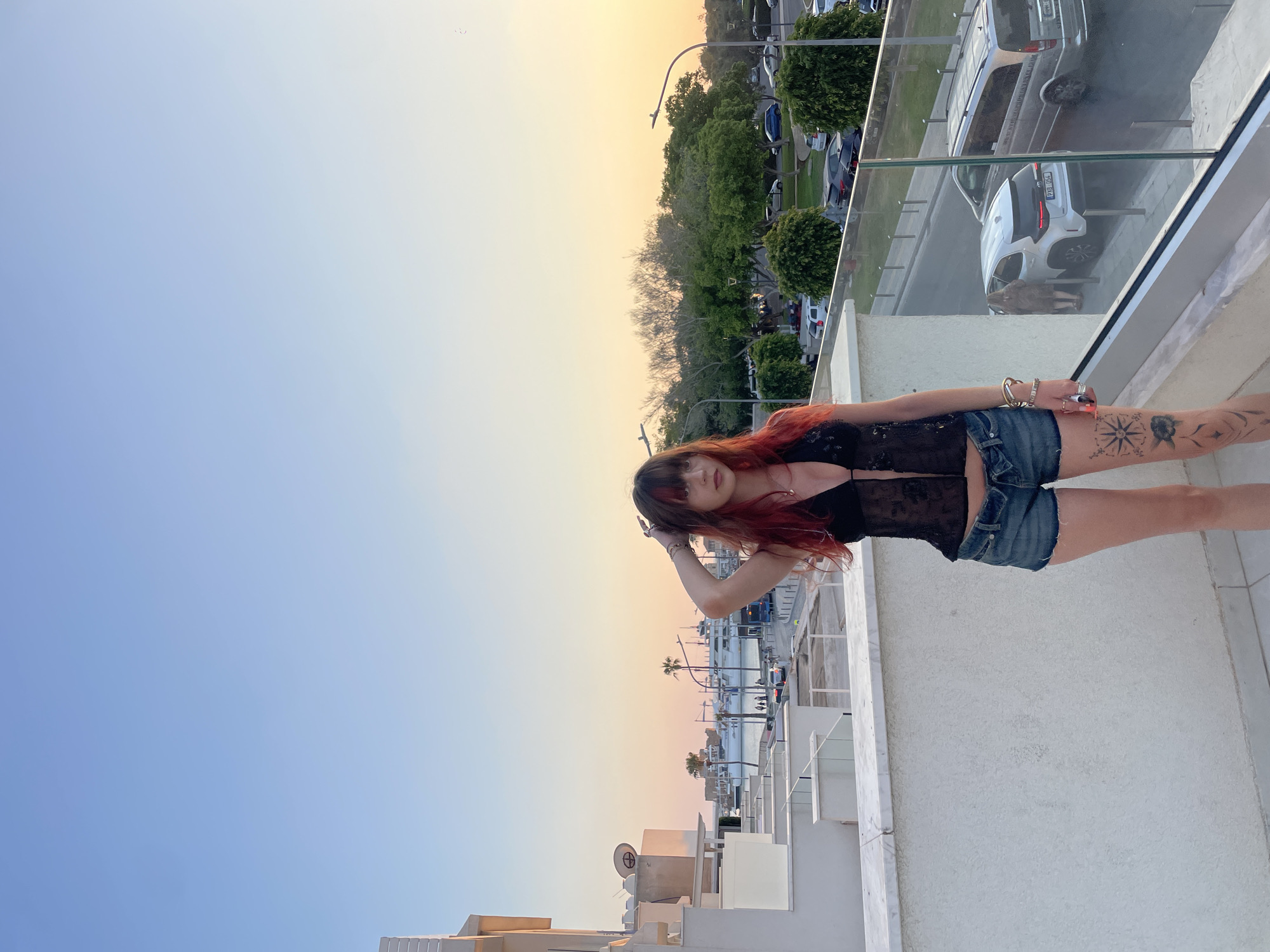 A young woman with long, dark hair with red streaks stands on a rooftop balcony in Paphos, Cyprus, at sunset. She is wearing a black lace top and denim shorts. Her left hand is resting on her head, and her right hand is down by her side. In the background, there is a harbor with boats and yachts, and a street with cars. The sky is a gradient of blue and orange, indicating the sun is setting. The architecture suggests a modern residential or tourist area.