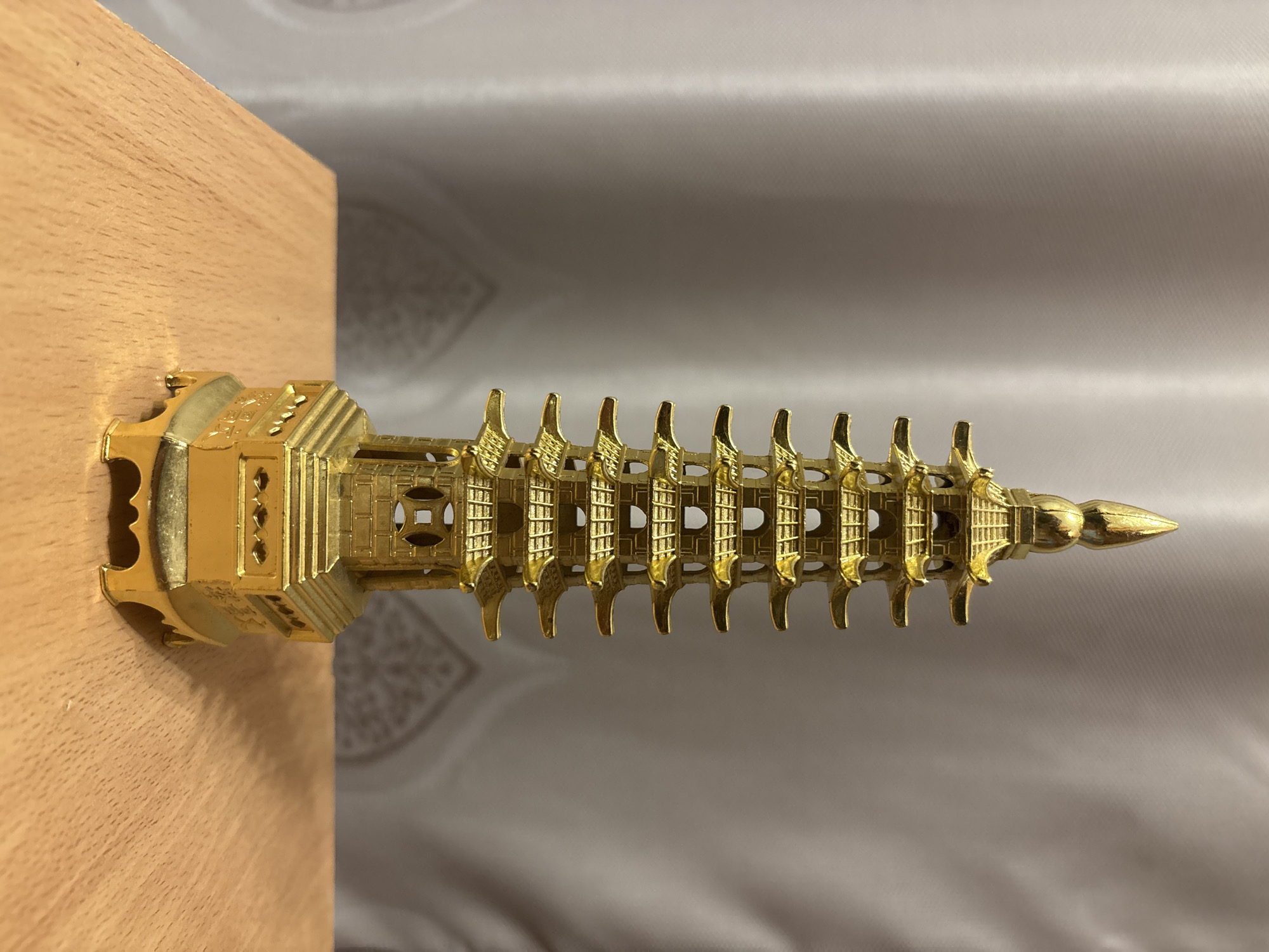 A golden, multi-tiered pagoda figurine rests on a light brown wooden surface, with a light grey curtain as a backdrop. The pagoda is intricately designed with repetitive architectural elements. The wooden surface is plain and serves as a base for the figurine. The curtain is smooth and slightly textured. The figurine is the primary subject of the image. There is no indication of the city of Zagatuy, Russia.