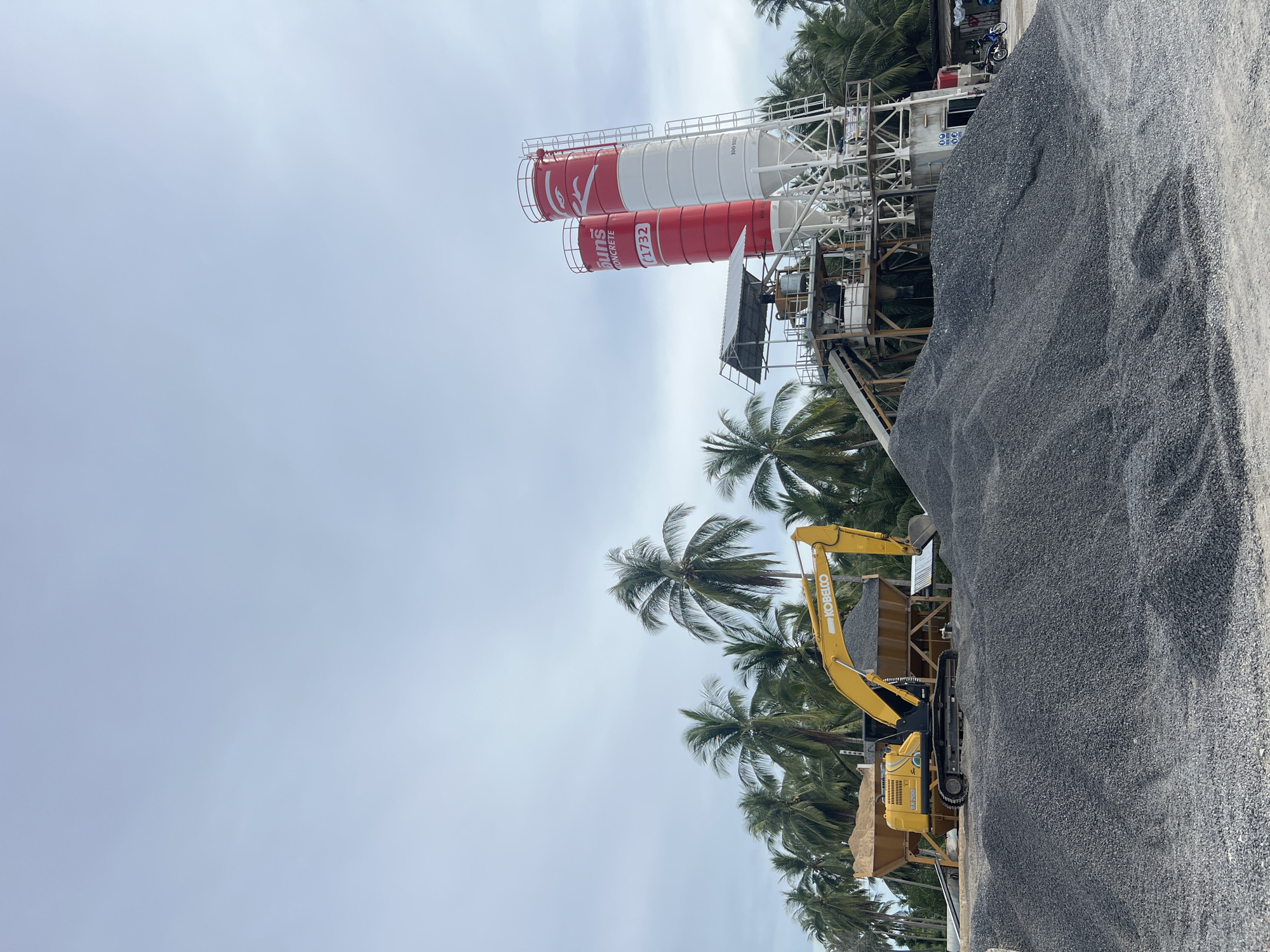 This image depicts an outdoor industrial scene, likely a construction site or quarry, situated amidst a tropical environment with numerous palm trees visible. The primary subjects are large, cylindrical concrete silos, painted red and white, with visible branding that appears to read "Collins Concrete" and a numerical sequence "1732" and "100-3021". These silos are part of a larger structure that includes scaffolding and conveyor belts, suggesting a material processing or mixing operation.
In the foreground, a large pile of dark grey aggregate or gravel dominates the right side of the frame. A yellow excavator, identified by the "KOBELCO" logo on its arm, is positioned near the pile and a hopper, actively engaged in moving or loading the material. The presence of the excavator and the vast quantity of aggregate point to ongoing construction or material handling activities.
The setting is characterized by lush green palm trees, indicative of a tropical climate. The sky is overcast with a pale, diffused light, suggesting it might be daytime, possibly during a cloudy period or early morning/late afternoon. There are no people clearly visible in the image. The overall mood is industrial and functional, with a backdrop of natural tropical scenery. The location context provided places the scene in Koh Phangan Subdistrict, Thailand, which aligns with the visual cues of palm trees and a tropical setting.