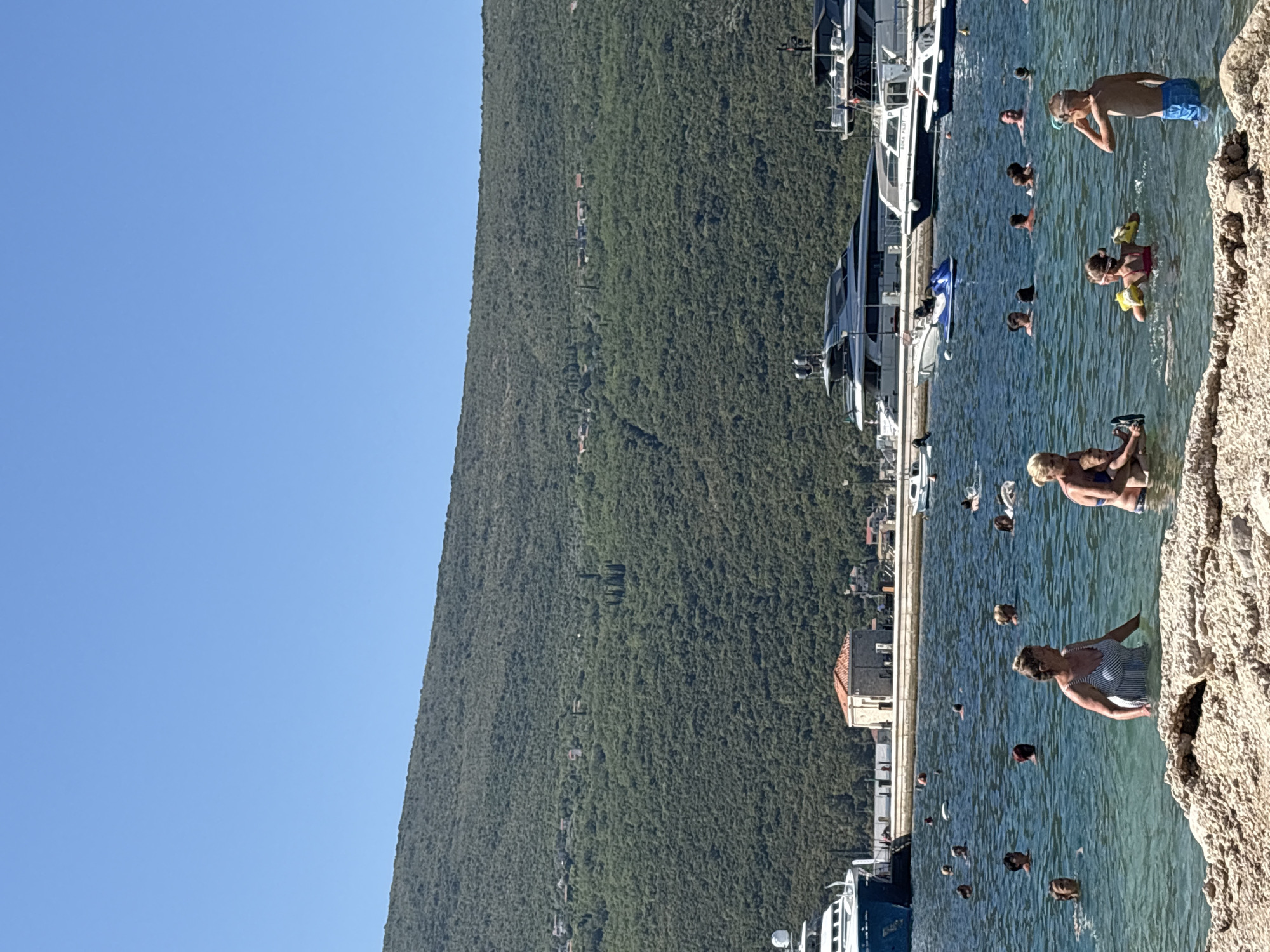 A wide-angle, eye-level shot captures a sunny day at a beach in Đenovići, Montenegro, with a backdrop of a densely forested, steep mountainside. The clear blue sky dominates the upper portion of the image. In the foreground, the edge of a rocky beach is visible, with clear, shallow turquoise water extending into the frame. The water is populated by numerous people of various ages, engaged in swimming and wading.
Several individuals are prominently featured in the mid-ground. Two women, one in a striped swimsuit and another in a blue bikini, stand in the water. A young boy, wearing blue swimming trunks and goggles, stands facing right, with his right hand raised to his face. A girl in a pink swimsuit with yellow arm floaties is in the water, facing left. A woman with blonde hair holds a small child in her arms.
Further out in the water, many other people are swimming and interacting. In the background, a line of large, modern yachts are docked along a quay. Behind the yachts, a small, light-colored building with a red-tiled roof stands out, suggesting a waterfront establishment. The entire scene is bathed in bright sunlight, indicating a warm, summery day.