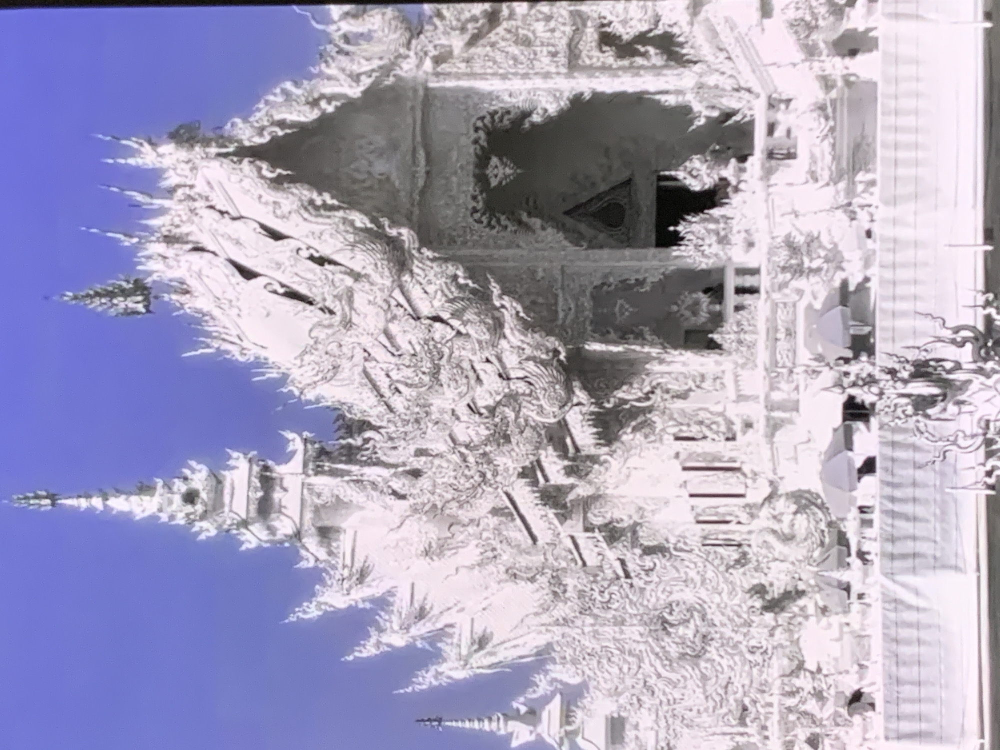 The image shows a white, ornate temple-like structure against a bright blue sky. The architecture features numerous spires, intricate carvings, and elaborate roof details. In the foreground, there is a white barrier, possibly a fence or wall, with decorative elements along its top. Several small umbrellas are visible behind the barrier, suggesting an outdoor area. A few indistinct figures can be seen within the temple complex. The overall impression is of a highly decorative and possibly religious building. The image is not located in Stavropol, Russia.